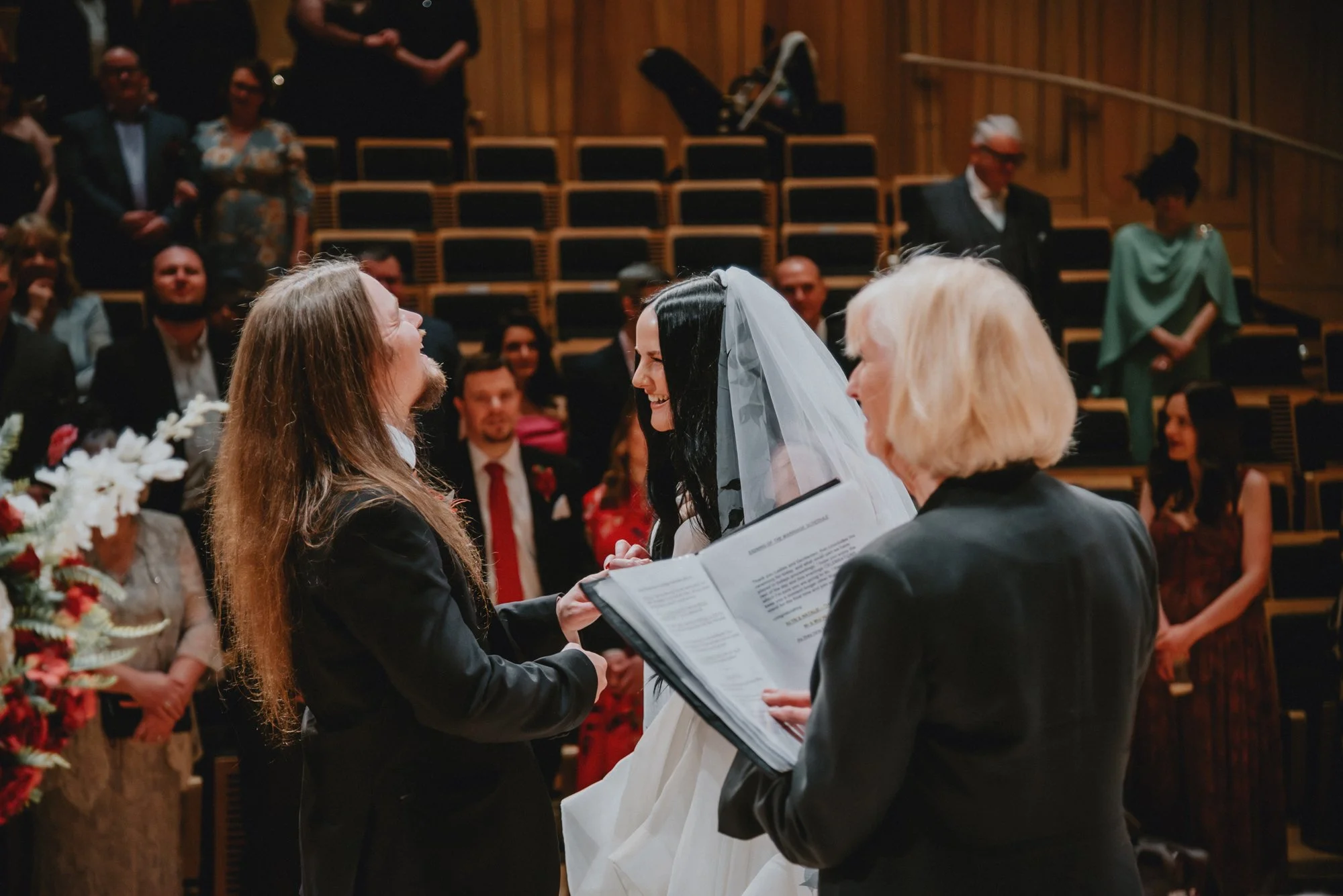 Bride and groom exchanging vows at their wedding ceremony, with officiant reading from a book, in an indoor venue with seated guests in the background.