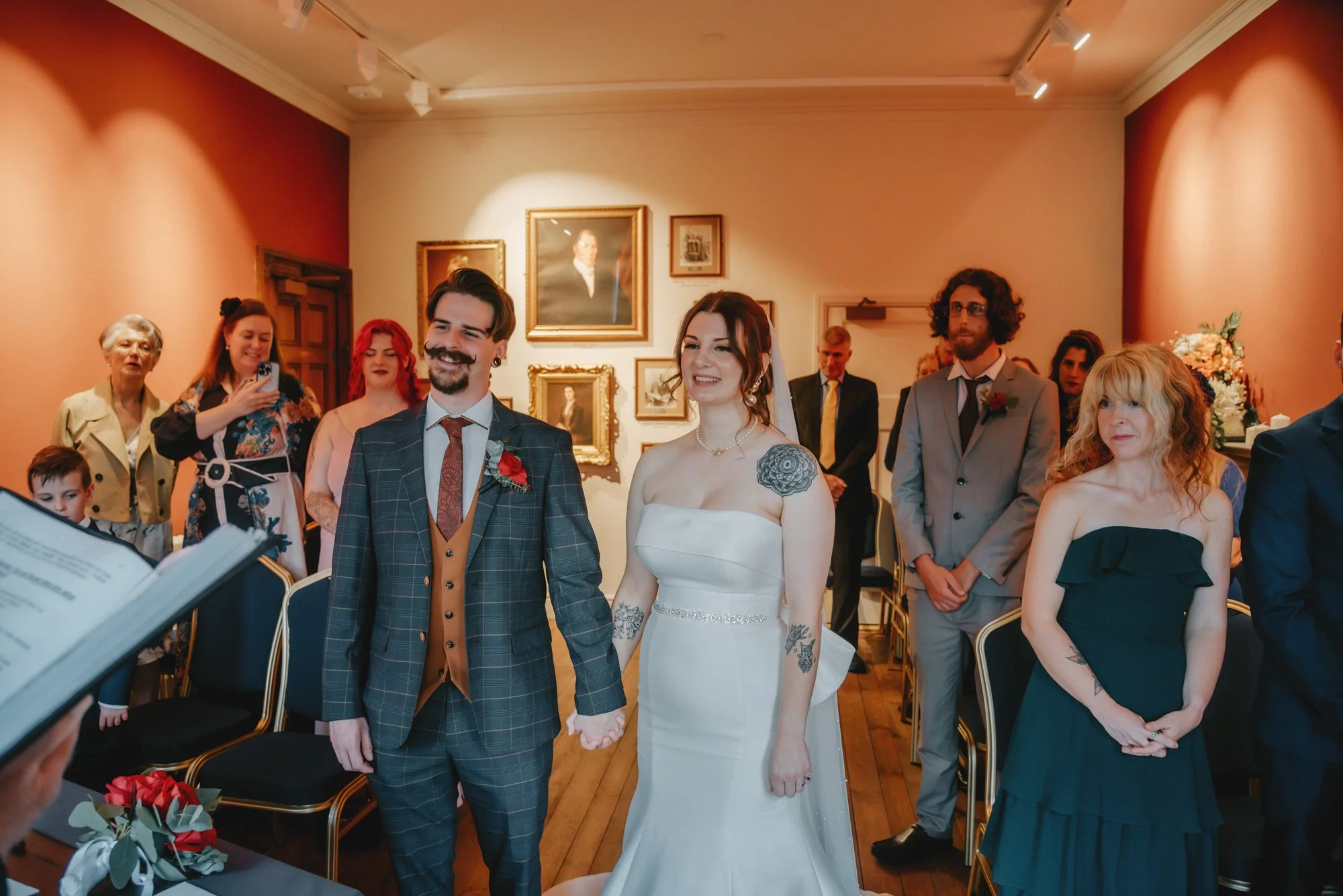 A couple getting married, holding hands, standing in front of friends and family in a decorated room, with framed artwork on the wall.
