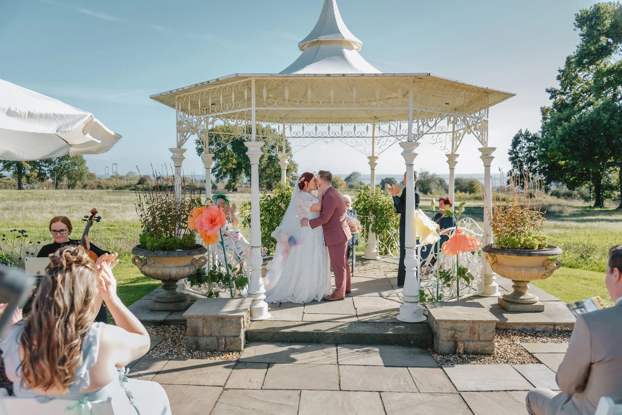 Bride and groom kissing under a white gazebo during their outdoor wedding ceremony, with friends and family clapping and taking pictures in a sunny field.
