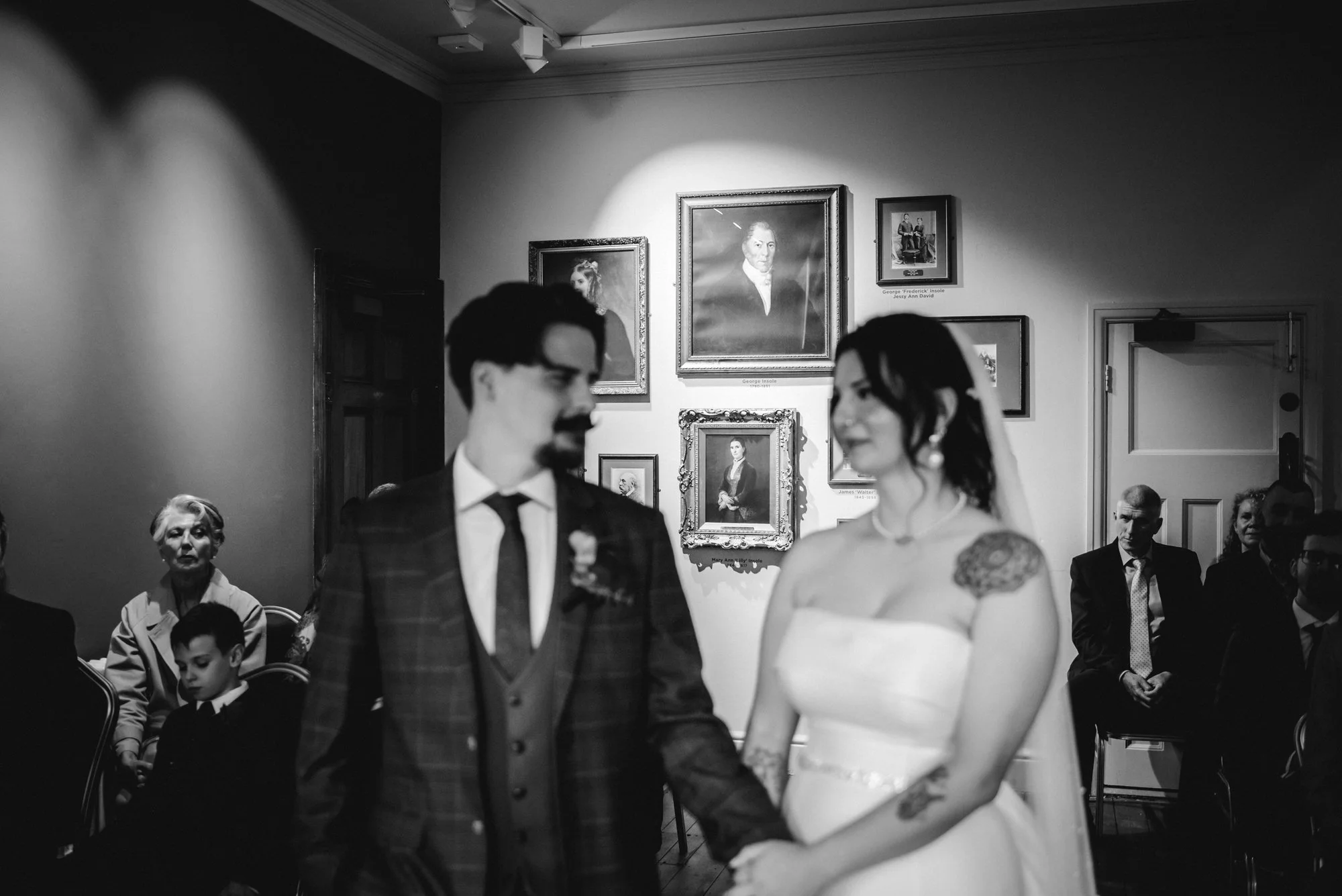 Black and white photo of a newly married couple holding hands during their wedding ceremony, with guests seated in the background and framed portraits on the wall.