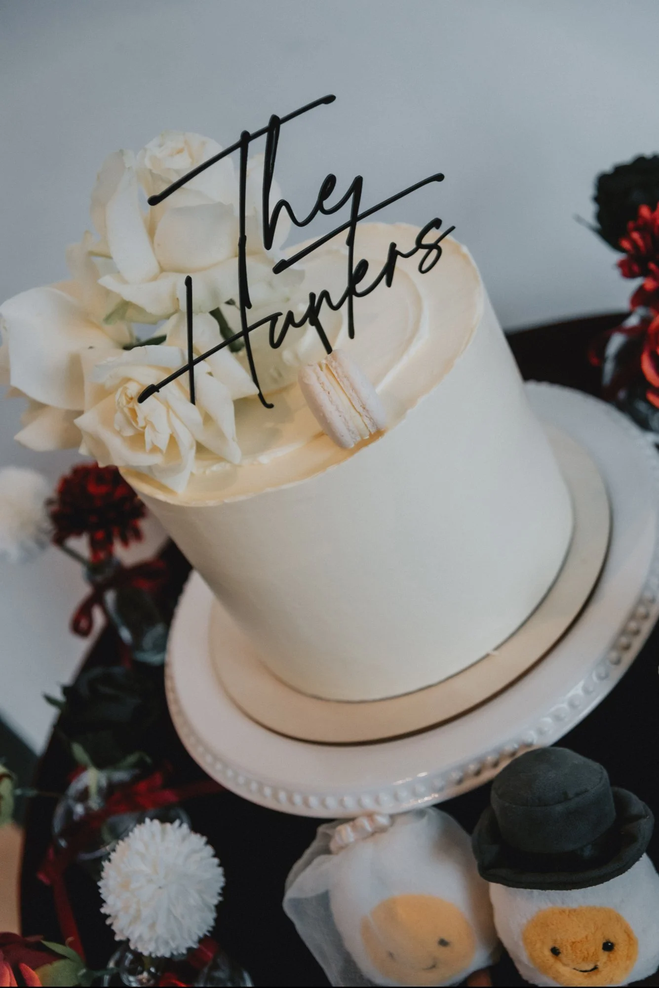 A white cake decorated with white flowers, a macaroon, and a topper that reads 'They Hapens'. The cake is on a white stand, surrounded by small decorative items and flowers.