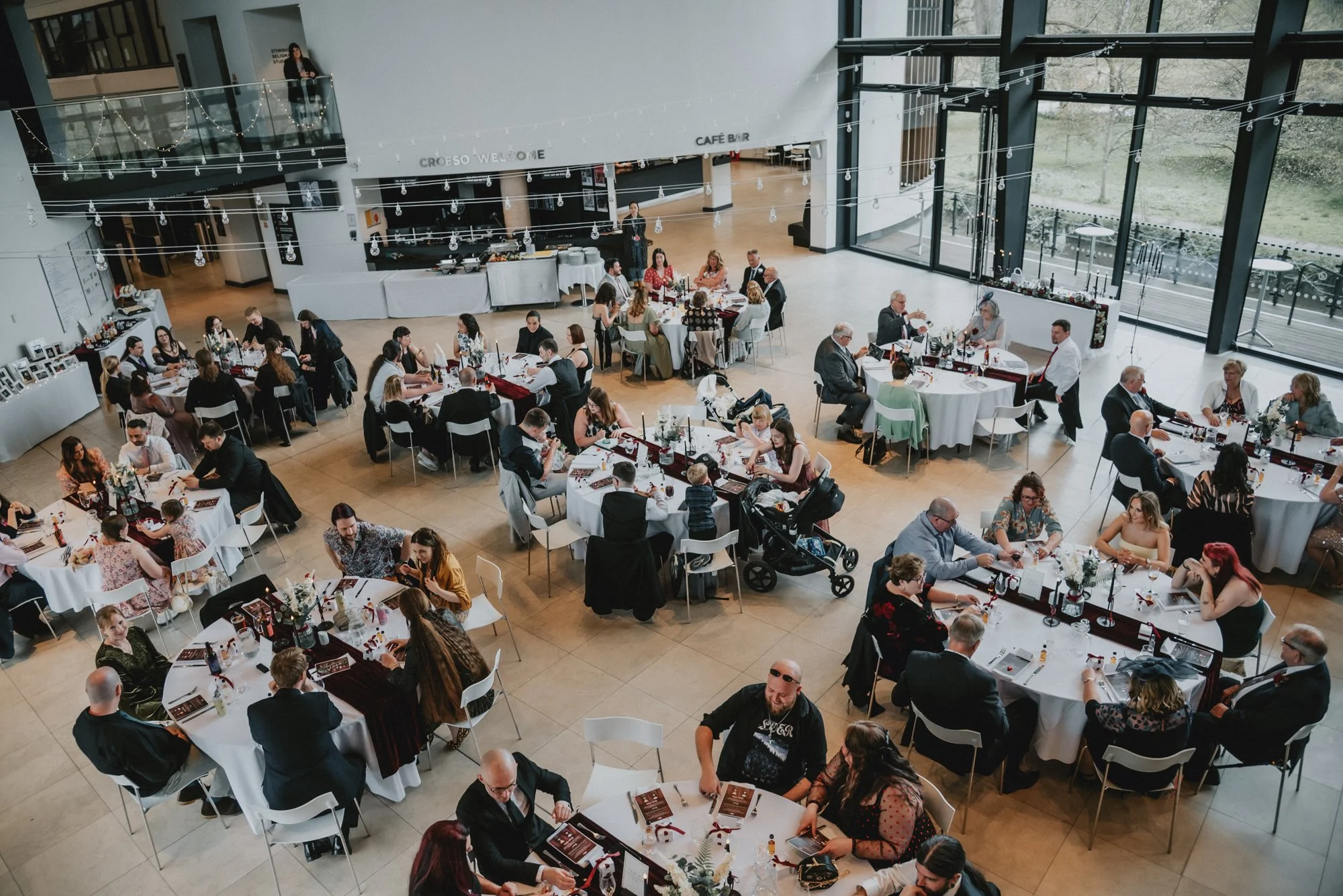 A large reception hall with multiple round tables set for a formal event. Guests are seated and engaging in conversation. The hall features large windows, modern decor, string lights, and a buffet area in the background.