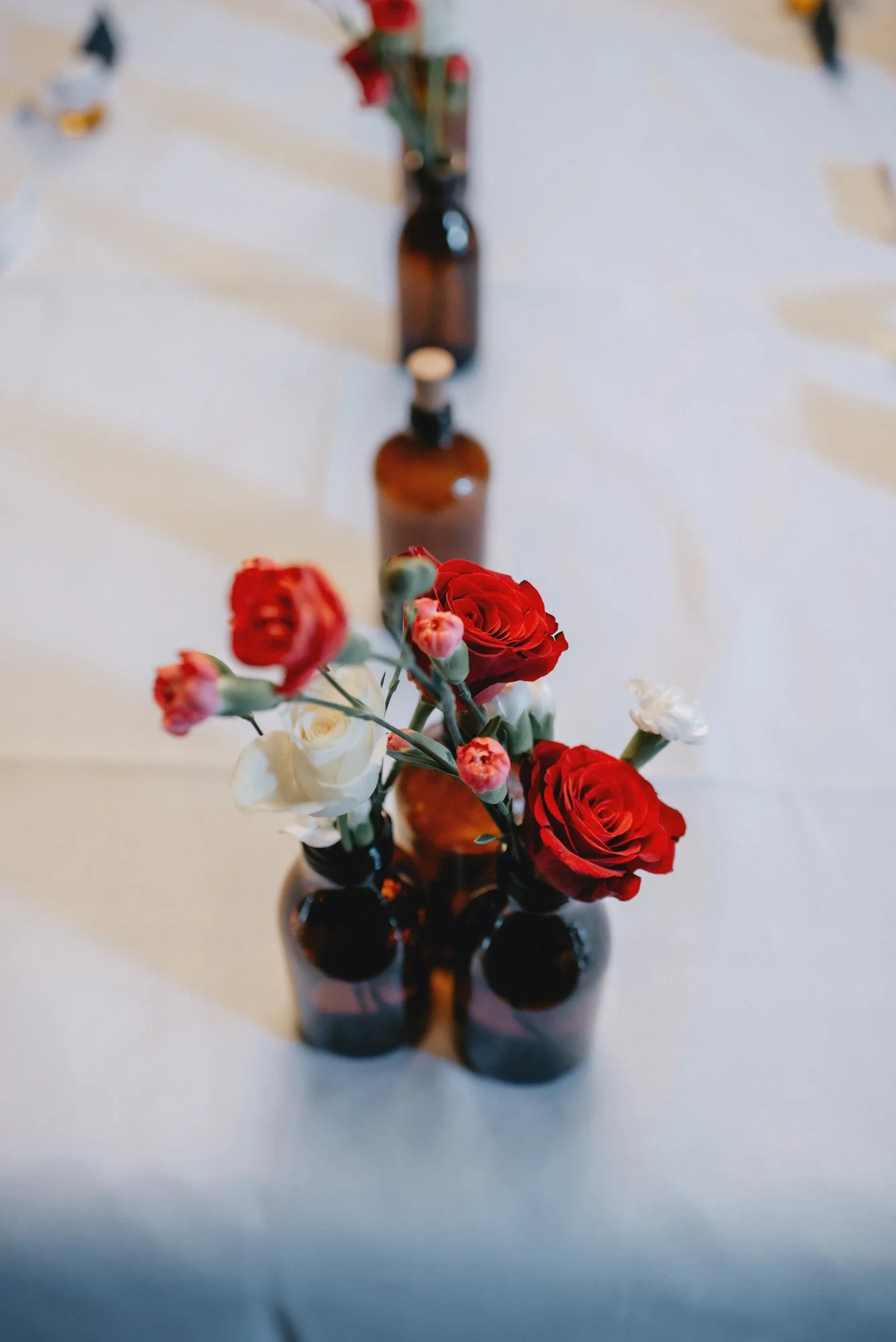 A top view of a table with small brown bottles holding red and white flowers arranged in a line as a centerpiece.