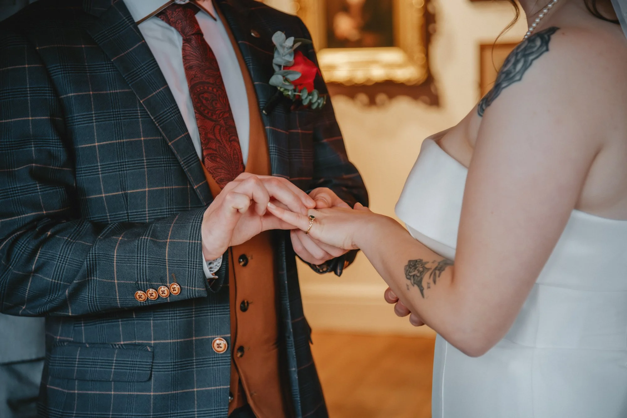 A couple exchanging rings during their wedding ceremony. The groom wears a dark plaid suit with a red tie and boutonniere, while the bride wears an off-shoulder white dress with tattoos on her arm.