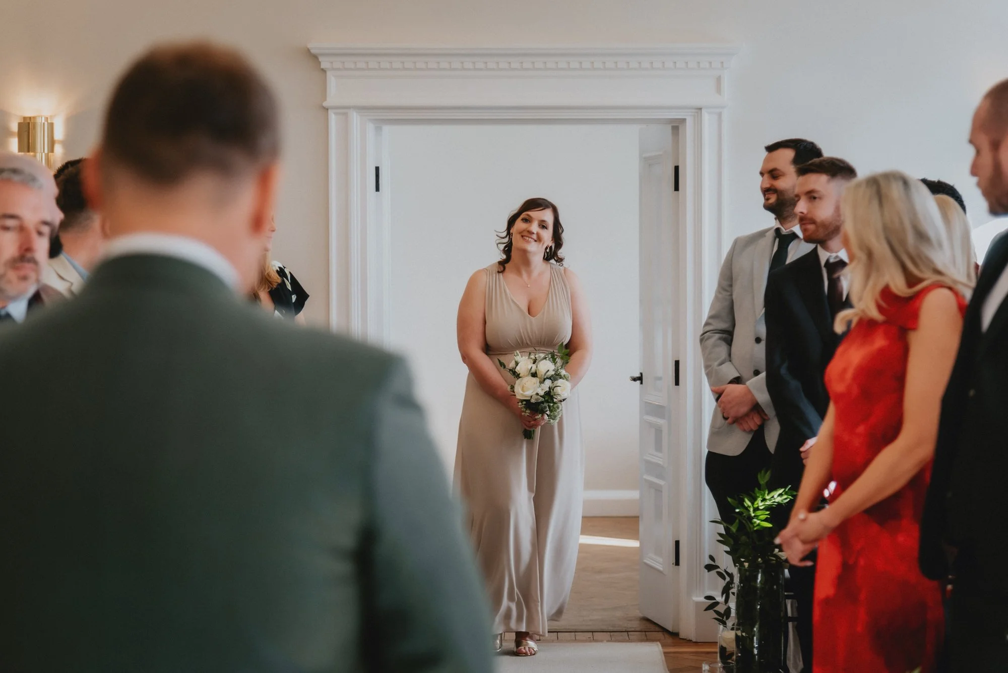 A woman in a beige dress holding a bouquet of white roses is smiling as she enters a room during a wedding ceremony, with people standing on either side.