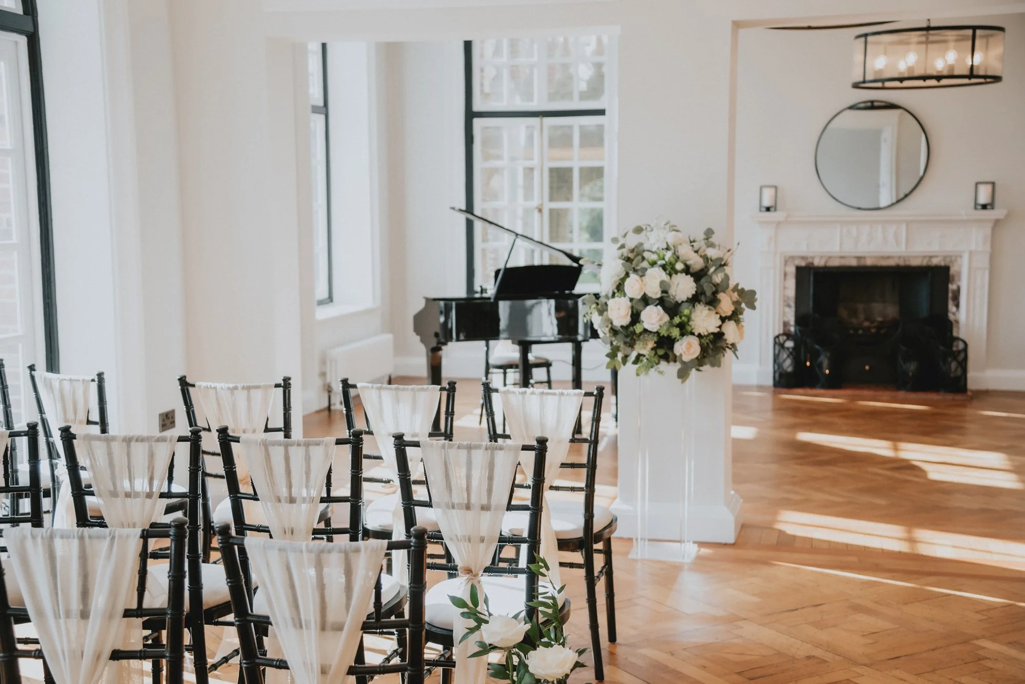 Elegant event room with rows of black chairs with white fabric drapes, a large floral arrangement on a stand, a grand piano, and a fireplace with a mirror and candles, prepared for a ceremony or celebration.