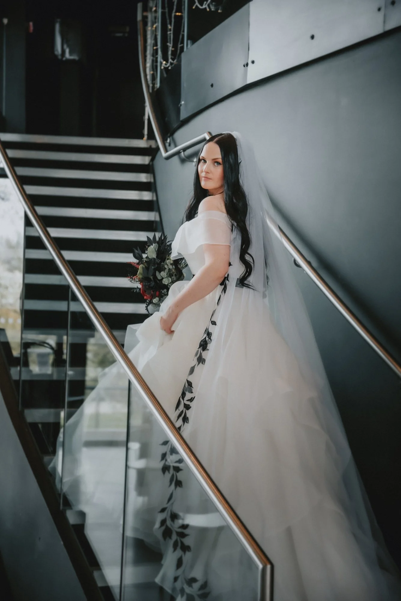 A woman in a white wedding dress with long dark hair and a veil, holding a bouquet, standing on a staircase with modern black and glass railing.