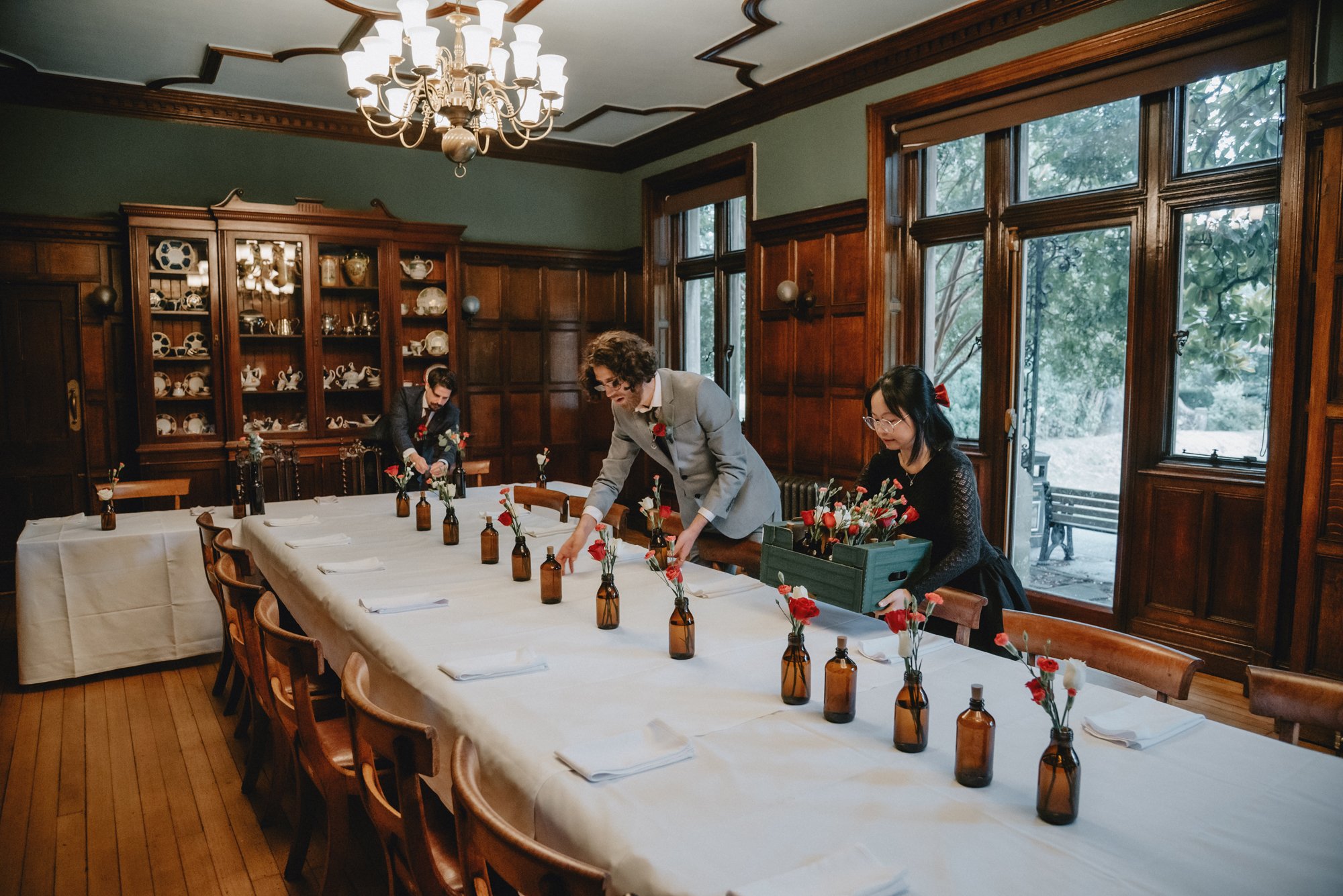 People setting up a long dining table with flowers in small glass bottles in a wood-paneled room with large windows.