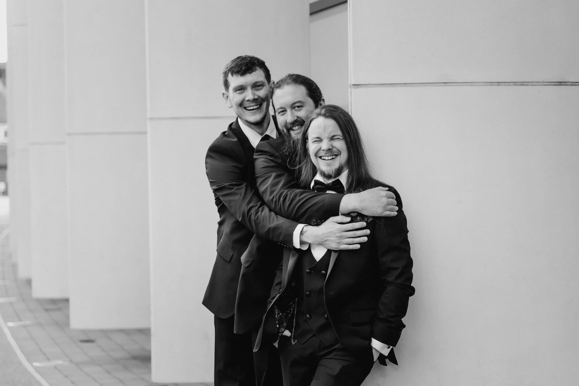 Three men in tuxedos smiling and hugging each other, standing outside near a building wall.