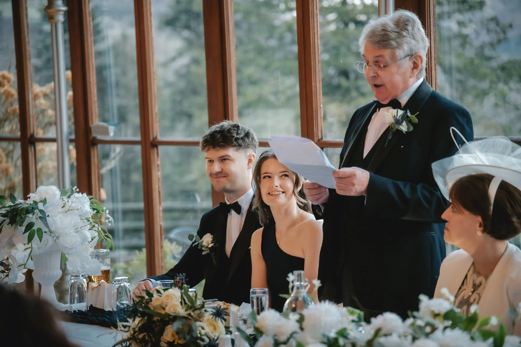 A wedding reception with a man in a tuxedo giving a speech, seated guests including a woman in a black dress smiling, and a woman in a hat, with floral decorations on the table and large windows behind them.