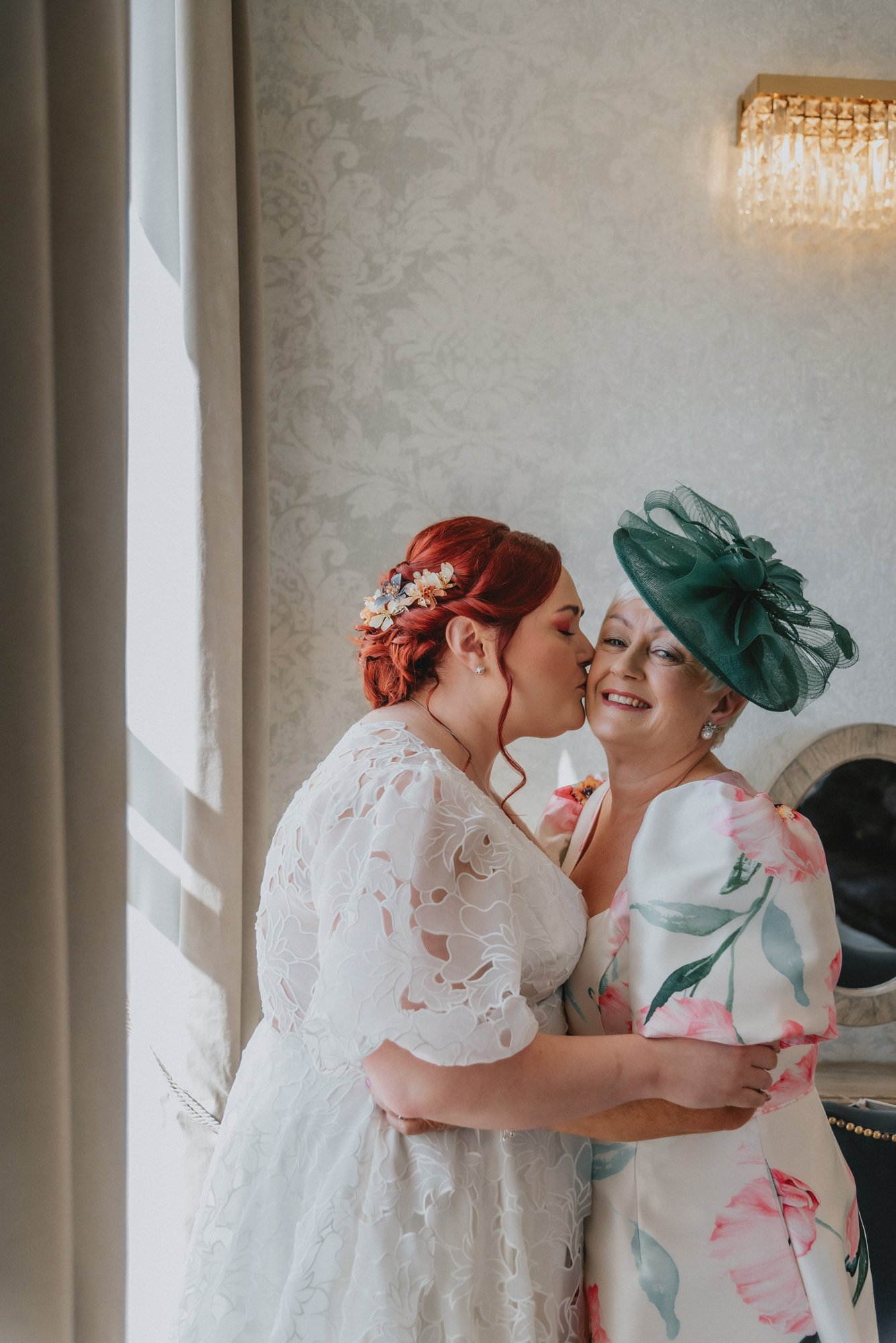 A bride with red hair and floral hair accessories kisses her smiling mother, who is wearing a green hat and floral dress, indoors near a window and chandelier.