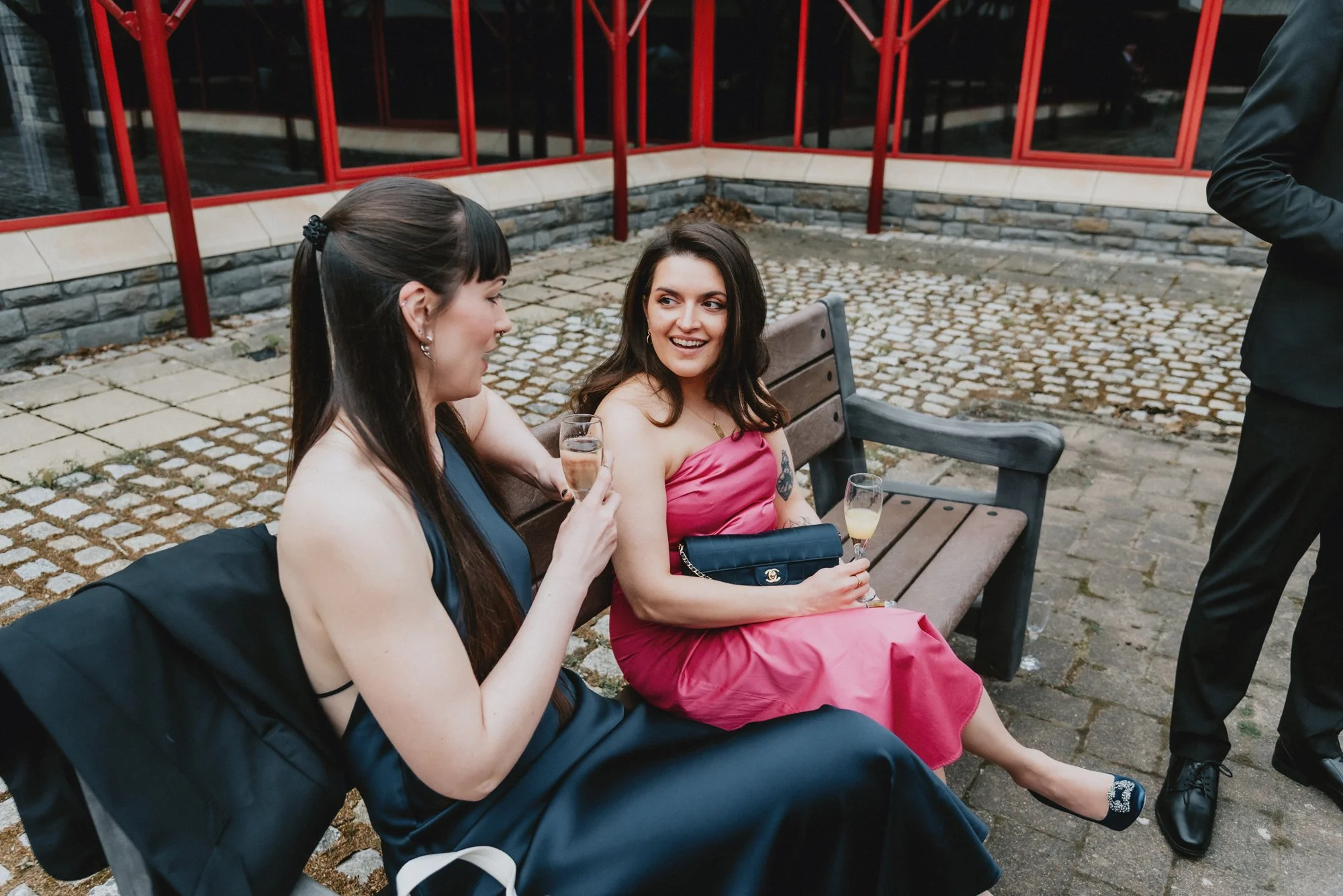 Two women in formal dresses sitting on a bench outdoors, holding glasses of champagne, and smiling at each other, with a man standing nearby.