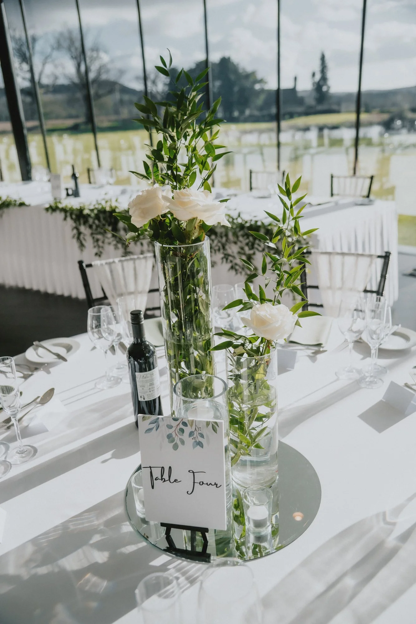Elegant wedding reception table with floral centerpieces, wine, and glassware, in a bright room with large windows showing outdoor scenery.