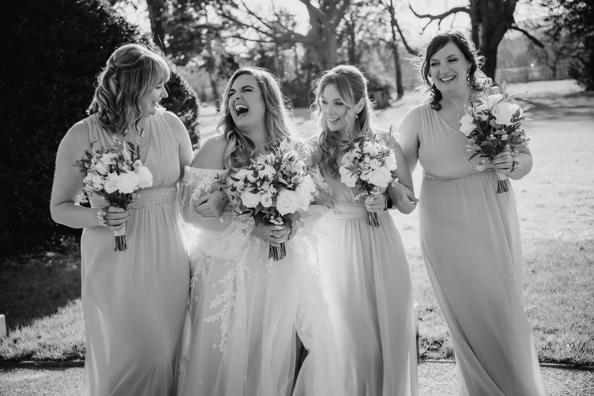 Black and white photo of a bride in a wedding dress and her four bridesmaids walking outdoors, holding bouquets and laughing