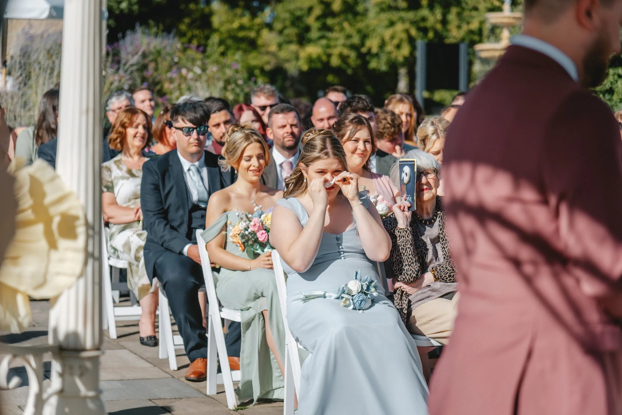 People attending an outdoor wedding ceremony, with a woman in a light blue dress crying and holding a tissue, in front of a man in a maroon suit.