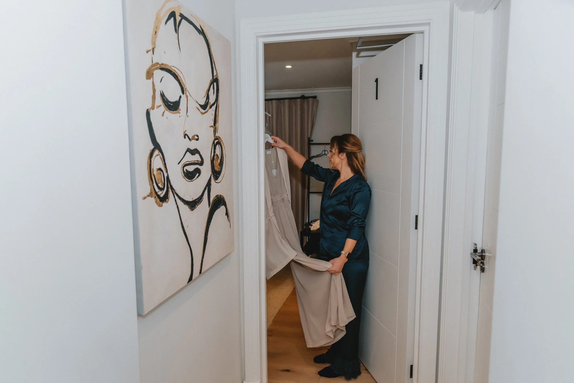 A woman in dark blue pajamas is standing in a doorway, holding a beige curtain with one hand and reaching towards a wall-mounted device with the other. The room behind her has beige curtains, a clothing rack, and a small table. On the white wall besi