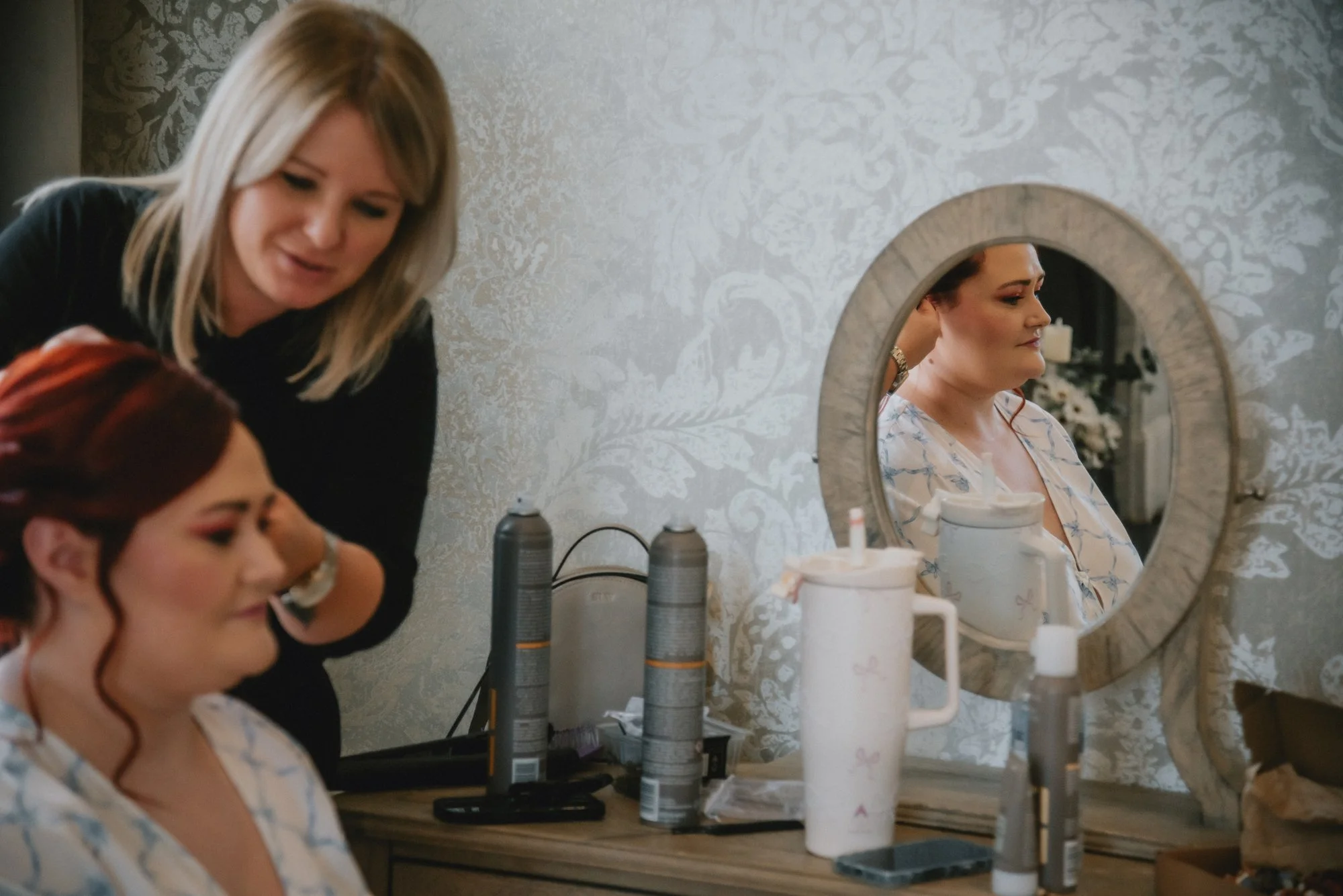 A woman with blonde hair and a black top is styling a woman's hair with red hair. The woman getting her hair styled is sitting in front of a mirror, wearing a light-colored robe with a floral pattern, and has a relaxed expression.