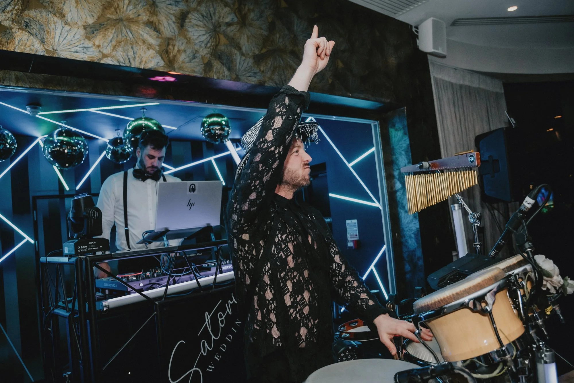A DJ in a white shirt and black suspenders is working on a laptop behind a table, with a man in a black lace top and a silver hat playing percussion instruments in the foreground at a wedding event, with dark walls and geometric blue lighting in the 