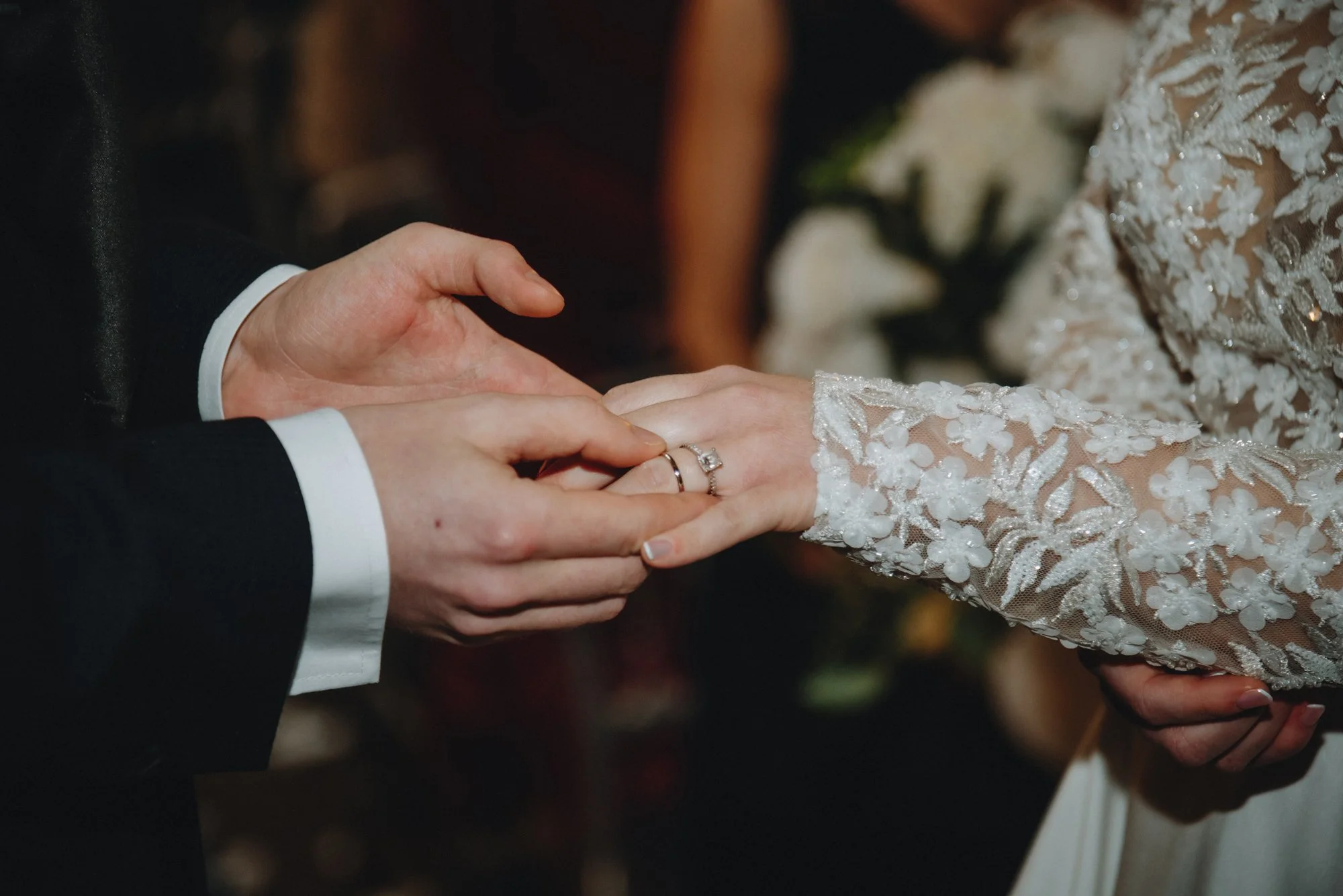 Close-up of a groom placing a ring on a bride's finger during a wedding ceremony. The bride is wearing a lace dress with floral embroidery, and the groom is dressed in a black suit with white shirt cuffs visible.