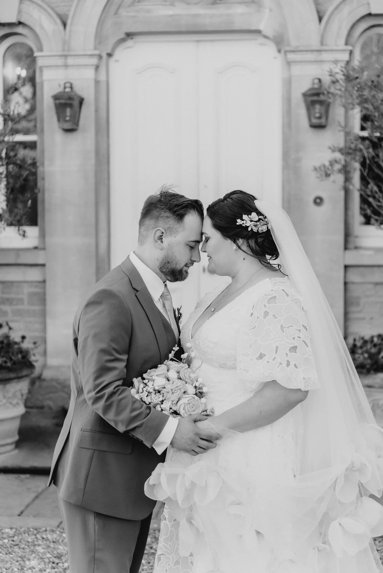 Black and white photo of a bride and groom standing close with foreheads touching, holding a bouquet of flowers, in front of a decorative door.