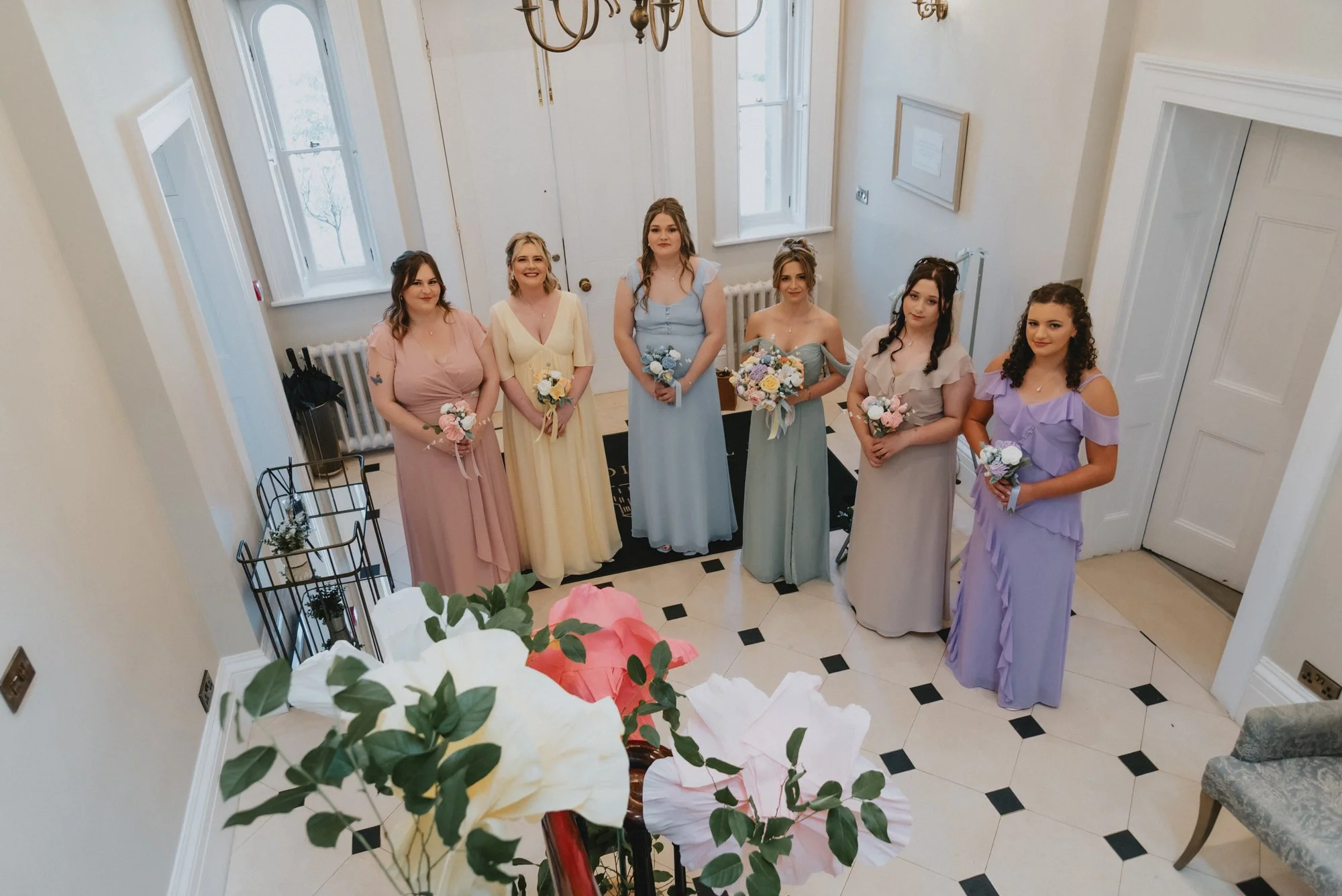 Six women dressed in pastel-colored dresses standing side by side indoors, holding flowers, in a bright room with a chandelier, windows, and black and white tiled floor.