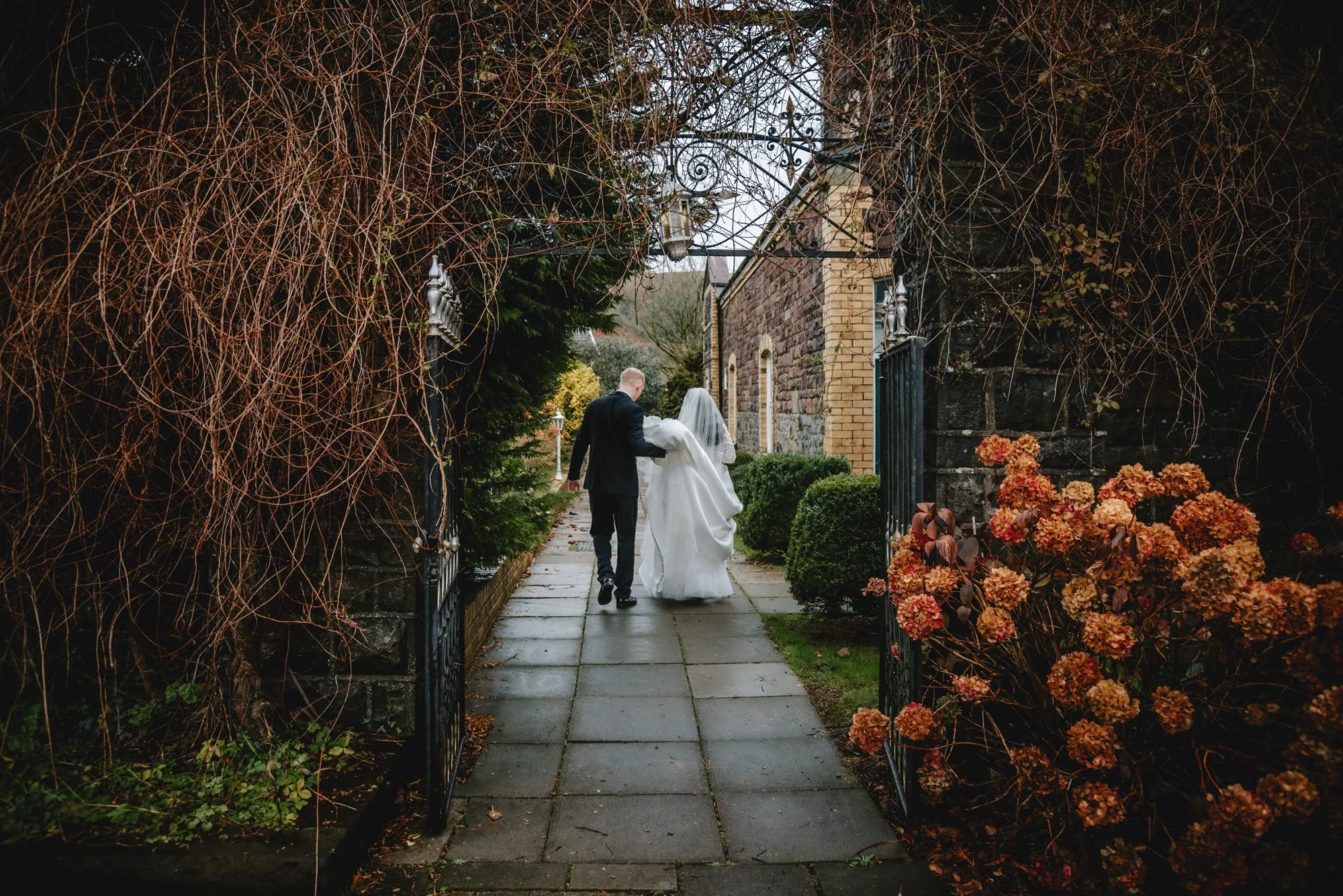 Bride and groom walking through a garden gate on their wedding day, with late autumn foliage and a stone building in the background.