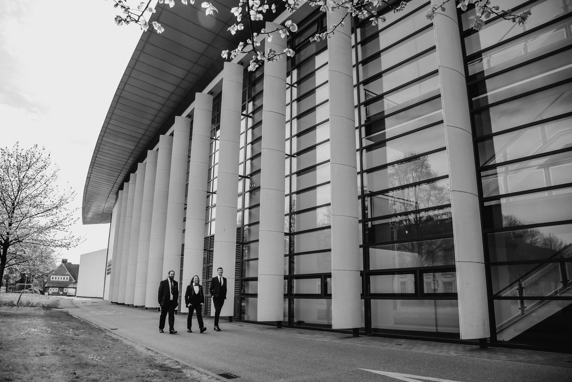 Three businesspeople walking along a sidewalk outside a modern building with large glass windows and tall white columns, with trees nearby.