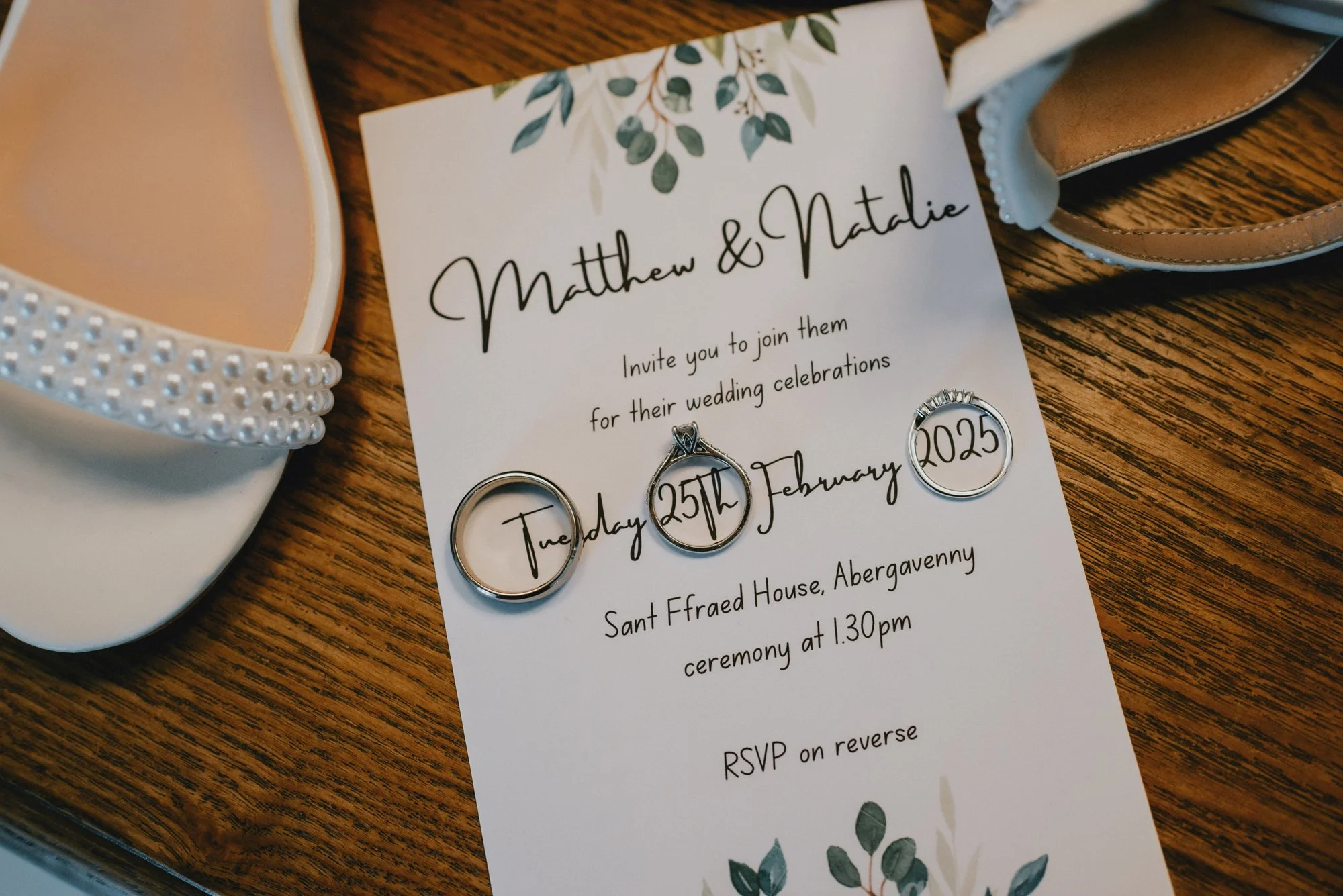 Wedding invitation for Matthew and Natalie with green leaf design, details including date, time, and location, with wedding rings and wedding shoes placed on a wooden surface.
