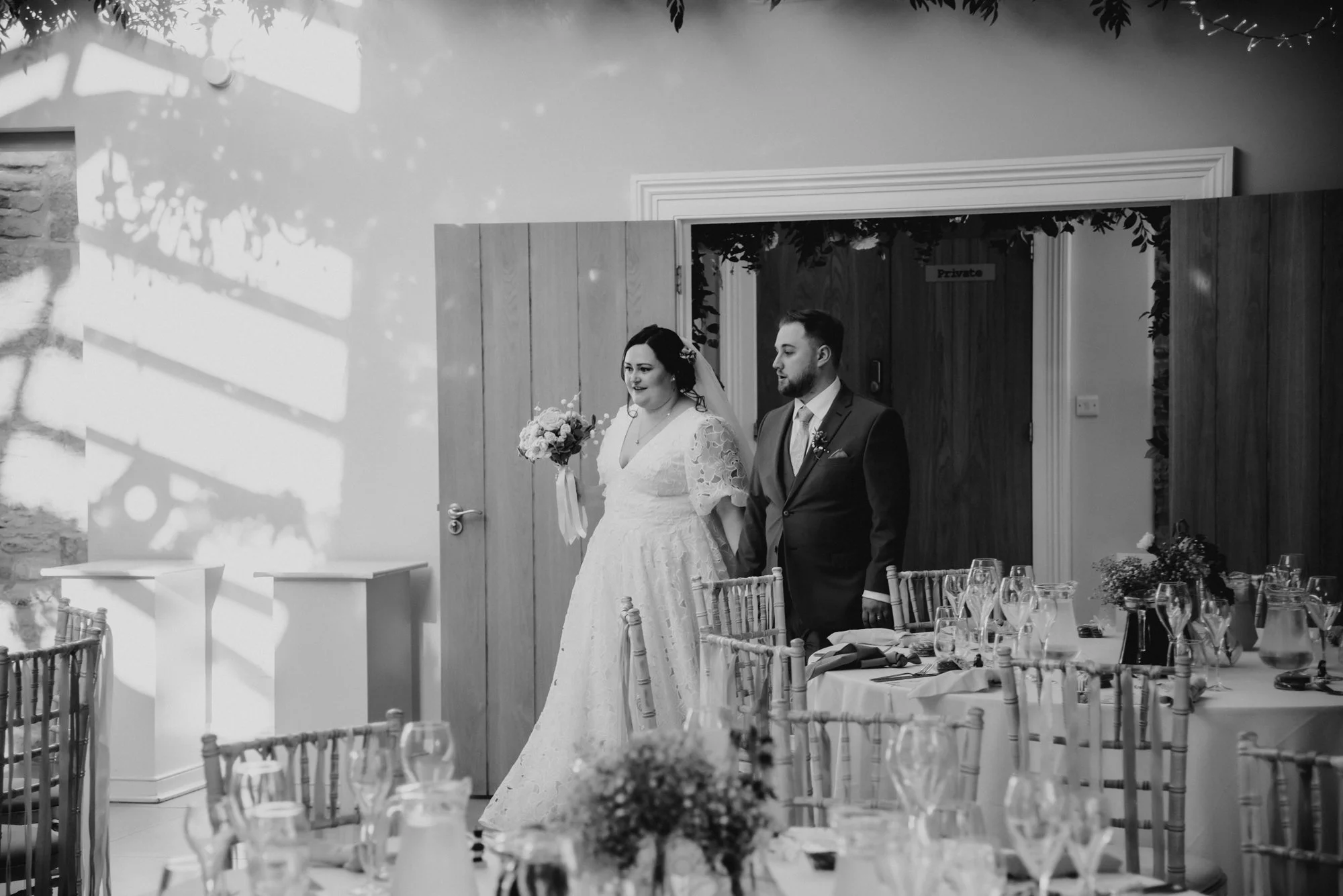 A bride and groom standing together in a wedding reception setting, photographer in black and white.