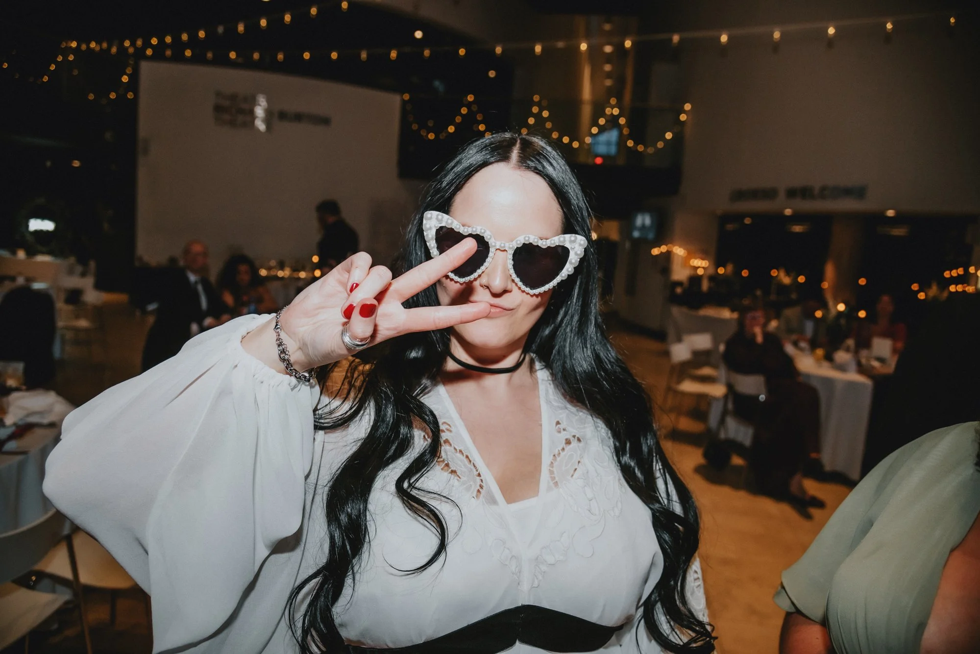 Woman with long black hair wearing white blouse and heart-shaped sunglasses making a peace sign at a party or celebration.