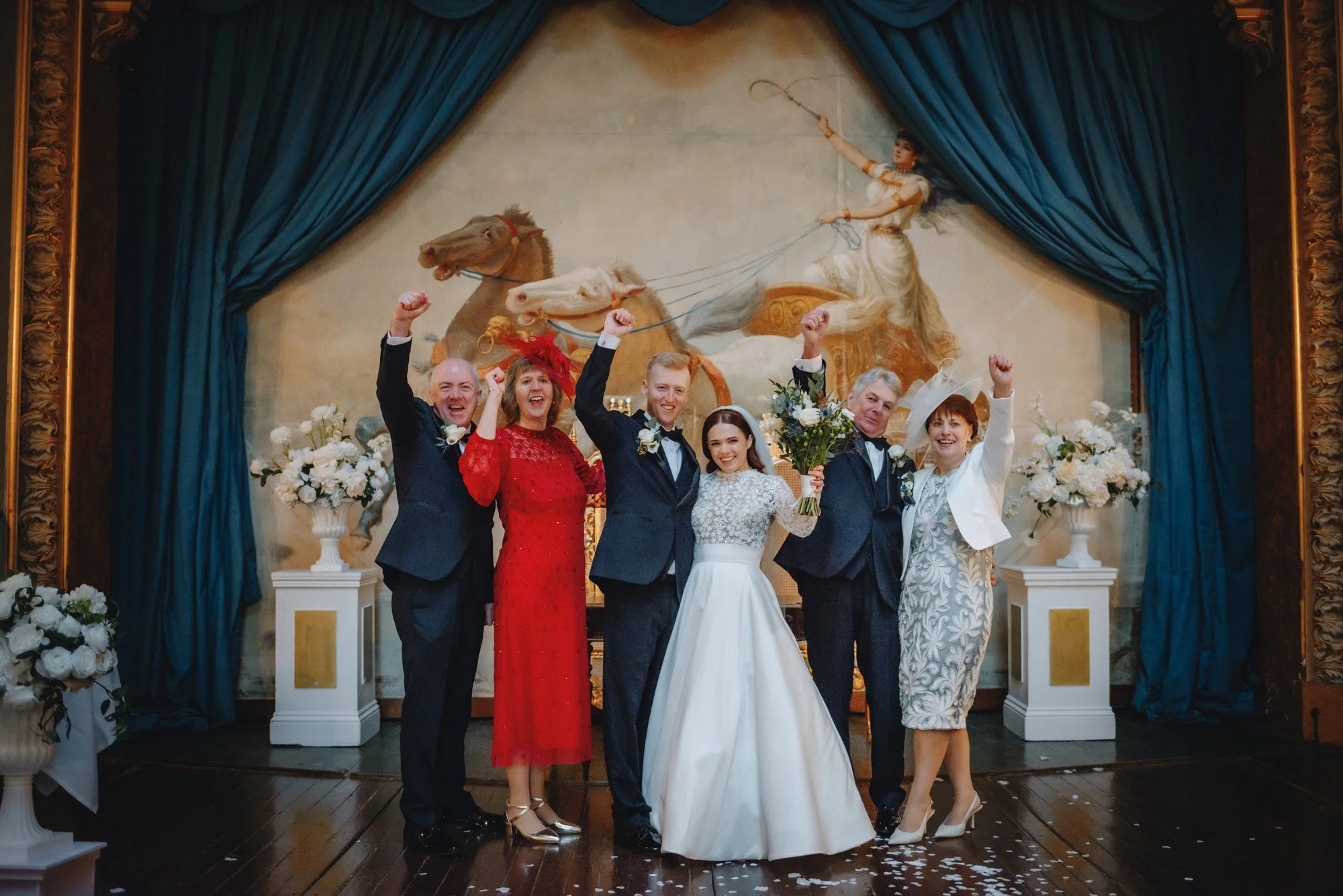 A wedding celebration with the bride and groom in the center, holding a bouquet, surrounded by family members on a decorated stage. The background features a large painting of a woman on a chariot pulled by horses, with blue curtains on either side.