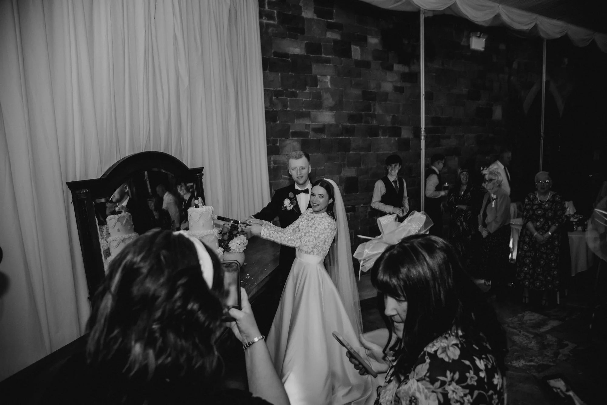 Black and white wedding photo of newlyweds cutting their wedding cake, surrounded by guests, in an indoor venue.