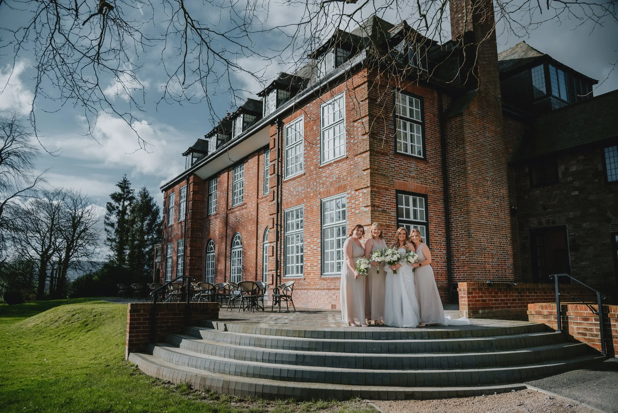 Four women dressed in white wedding gowns holding bouquets, standing on the steps in front of a large red brick building with tall windows, under a partly cloudy sky.