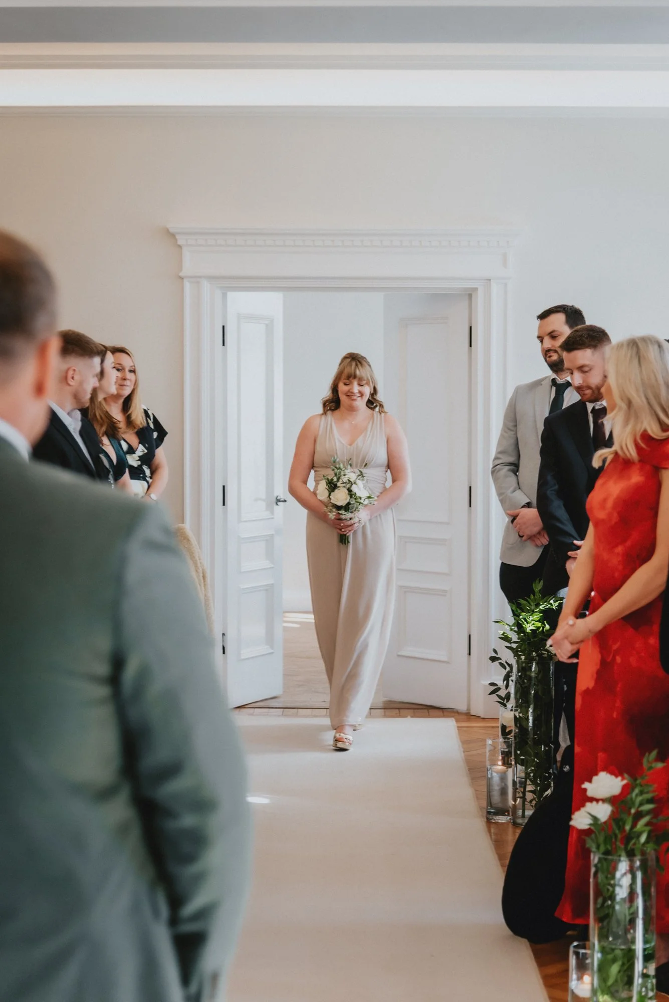A woman in a beige dress holding a bouquet walking down the aisle during a wedding ceremony with guests standing on either side.