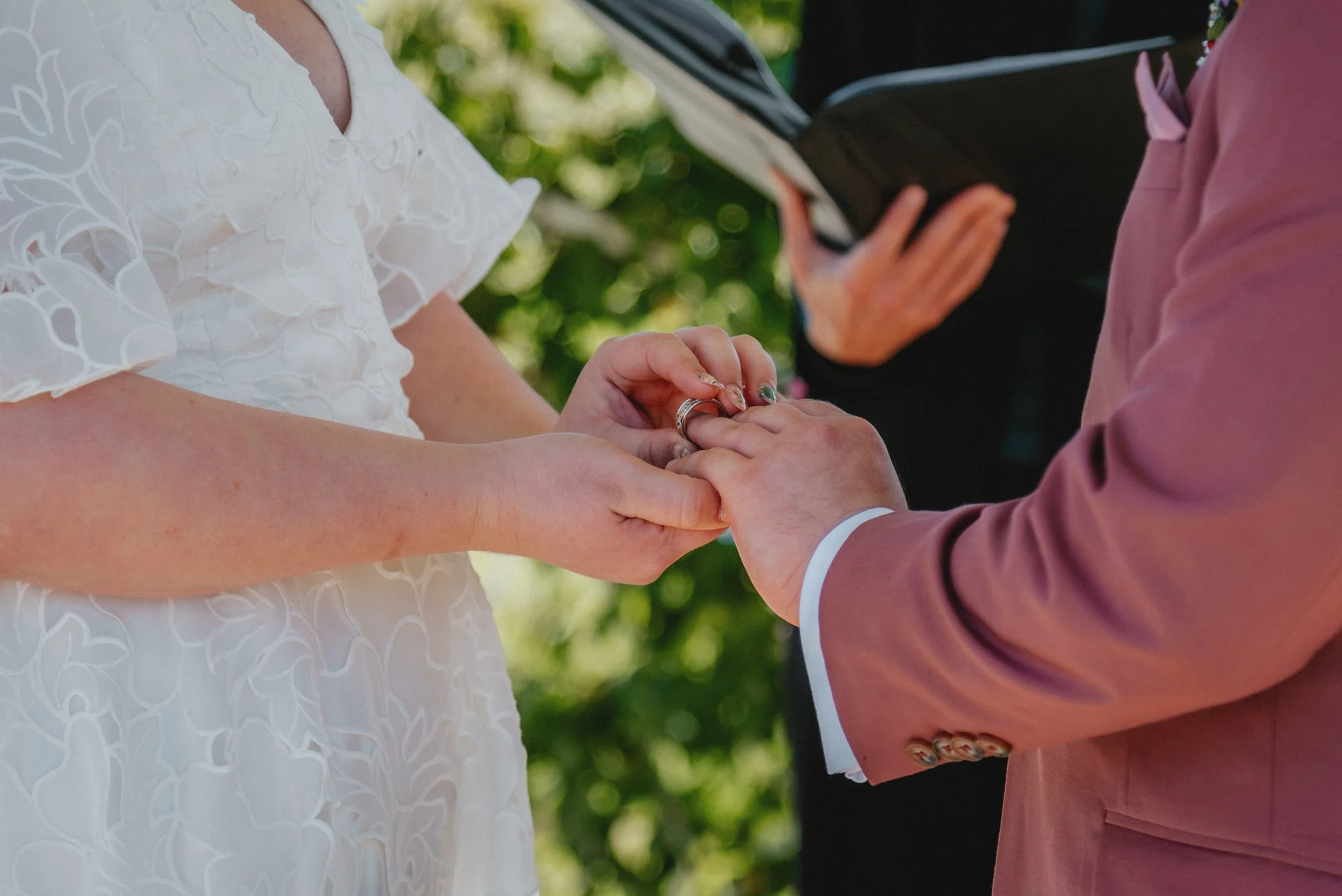 A priest officiating at a wedding ceremony, holding the hands of a bride and groom during the exchange of rings outdoors.