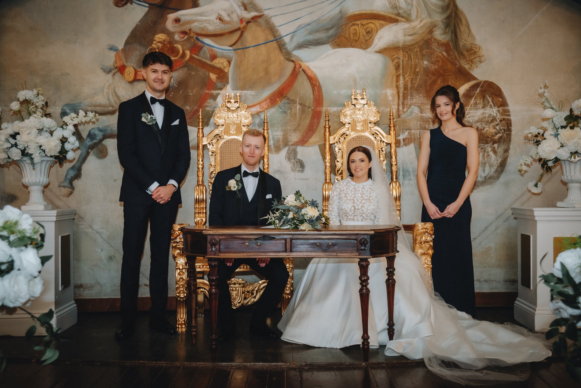 Wedding celebration with bride, groom, two attendants, and a person seated on a golden throne in front of a mural of horses and chariots, decorated with white flowers.