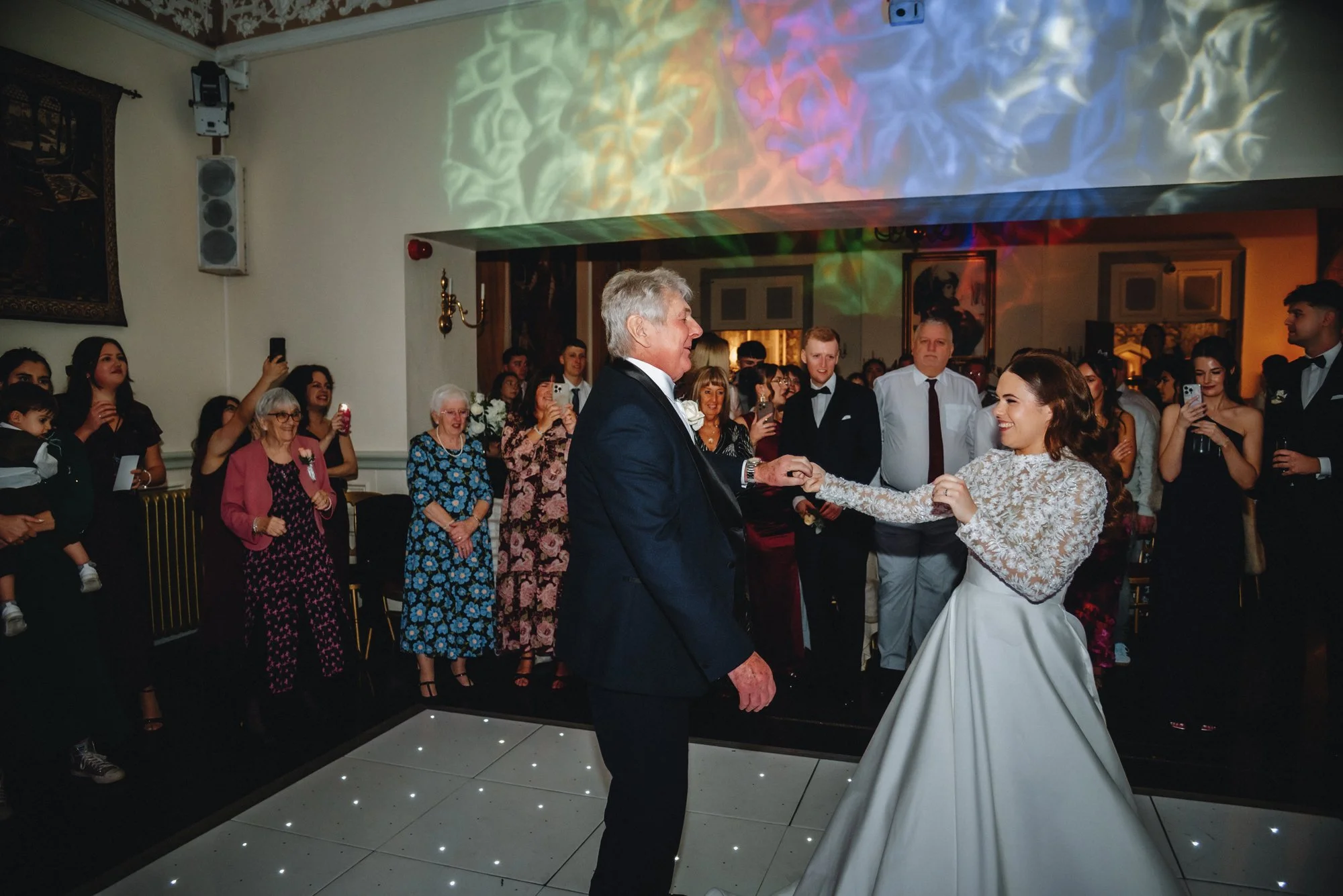 Older man and younger woman dancing at a wedding reception, surrounded by guests taking photos and watching.