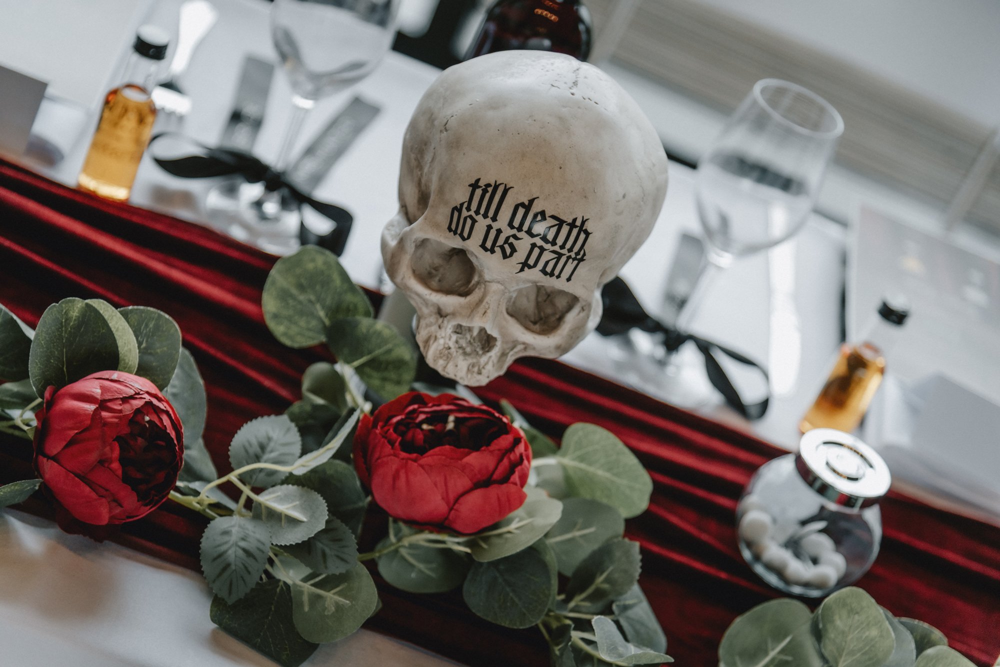 A skull replica with the phrase 'till death do us part' written on it, surrounded by red roses with green leaves, on a red velvet table runner. In the background, there are glasses, bottles, and a small jar.