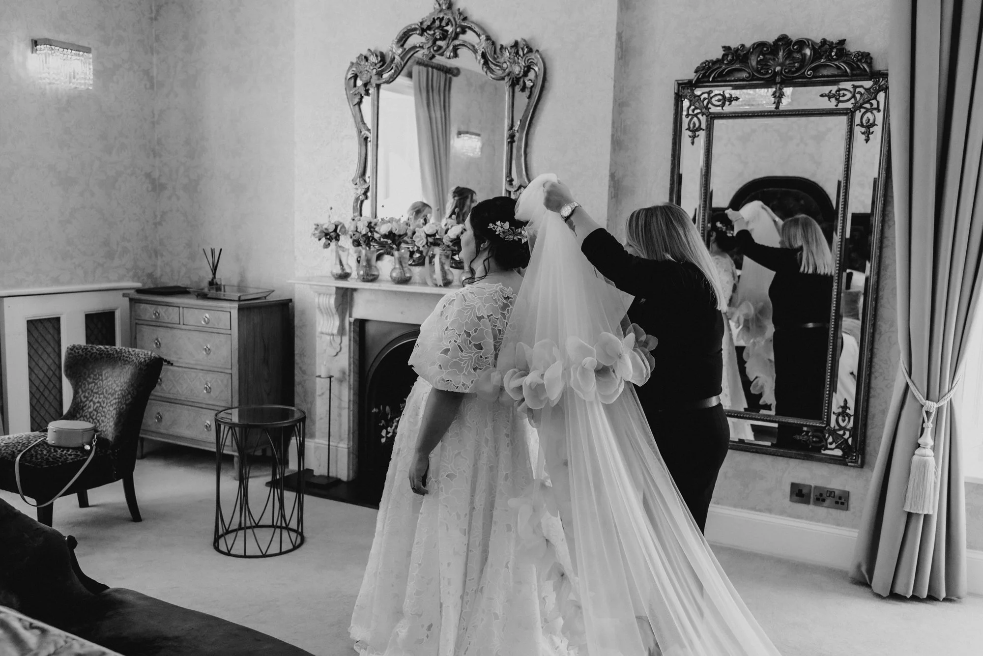 A bride with a veil and floral headpiece is helped by a woman in black with a veil, in a decorated room with large mirrors, floral arrangements, a fireplace, chairs, and curtains.