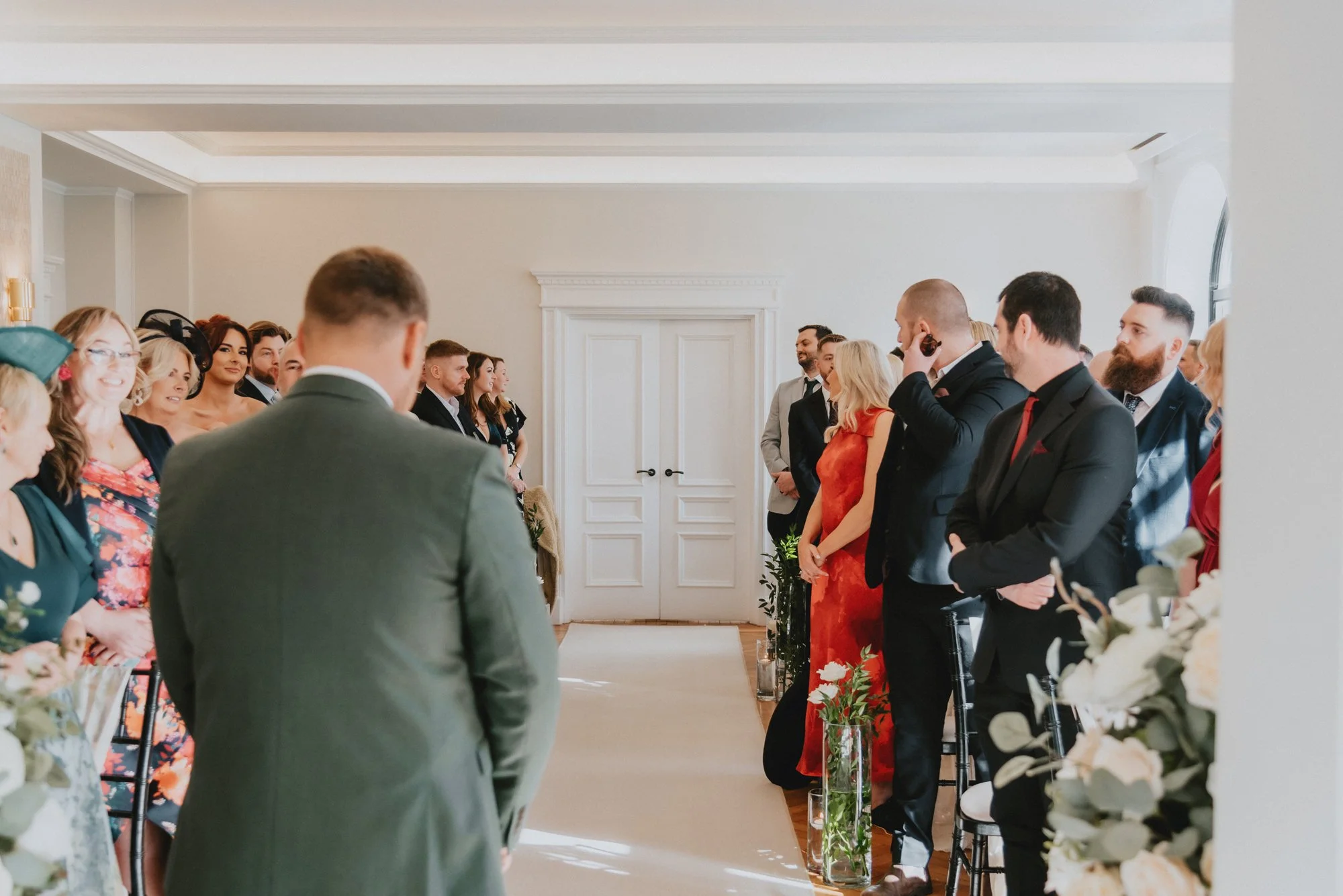 A wedding ceremony with guests standing on either side of the aisle, facing the couple. The bride and groom are not visible in the photo.