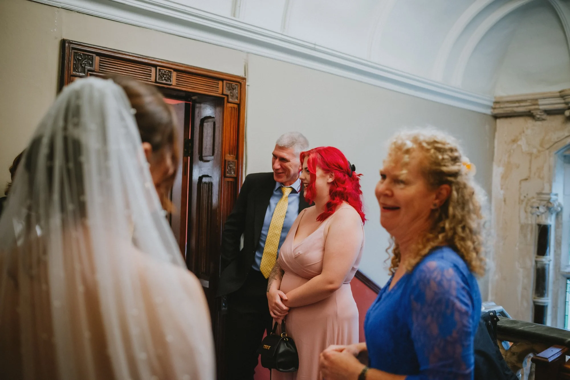 Group of people at a wedding, including a bride with a veil, a woman with red hair, a man in a suit, and another woman with curly blonde hair, in an elegant indoor setting.