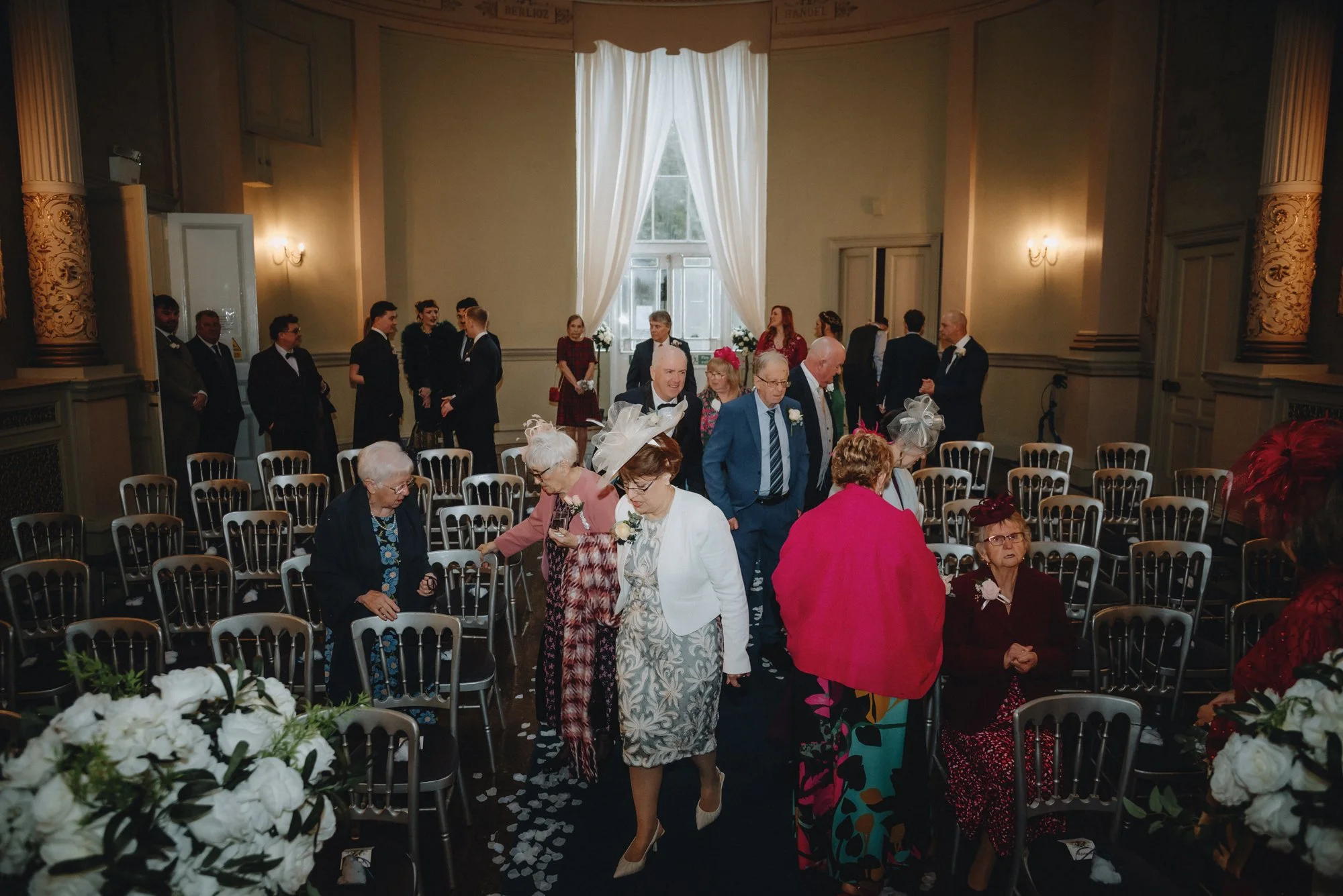 People gathered in a decorated room preparing for a wedding ceremony, with some elderly women and others standing or sitting, flower arrangements, and a large window with curtains in the background.