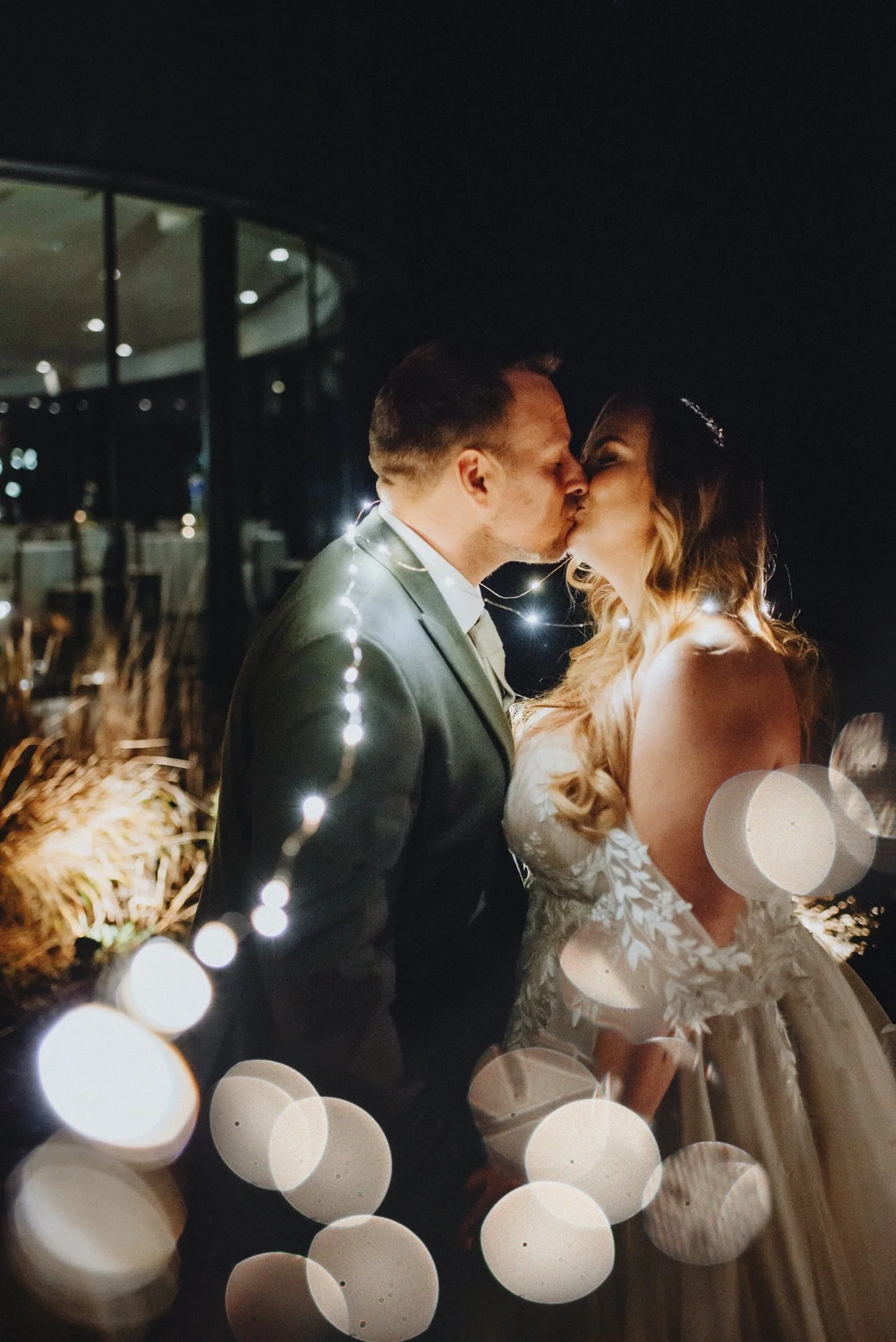 Couple in wedding attire exchanging a kiss at night, illuminated by string lights, with blurred bokeh effects in the foreground.