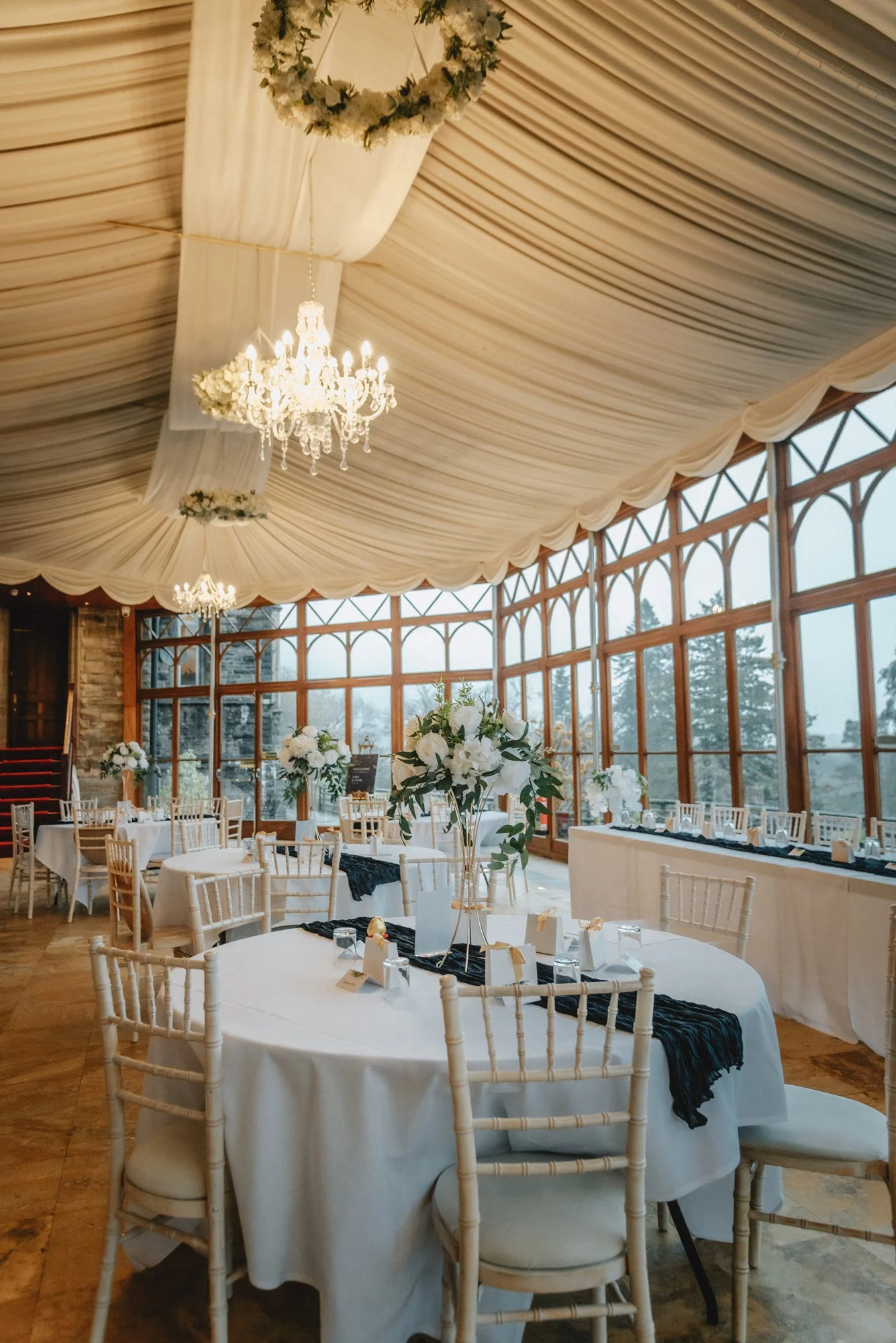 Elegant banquet hall with white tablecloths, gold chairs, floral centerpieces, and chandeliers, with large windows showing an outdoor view.