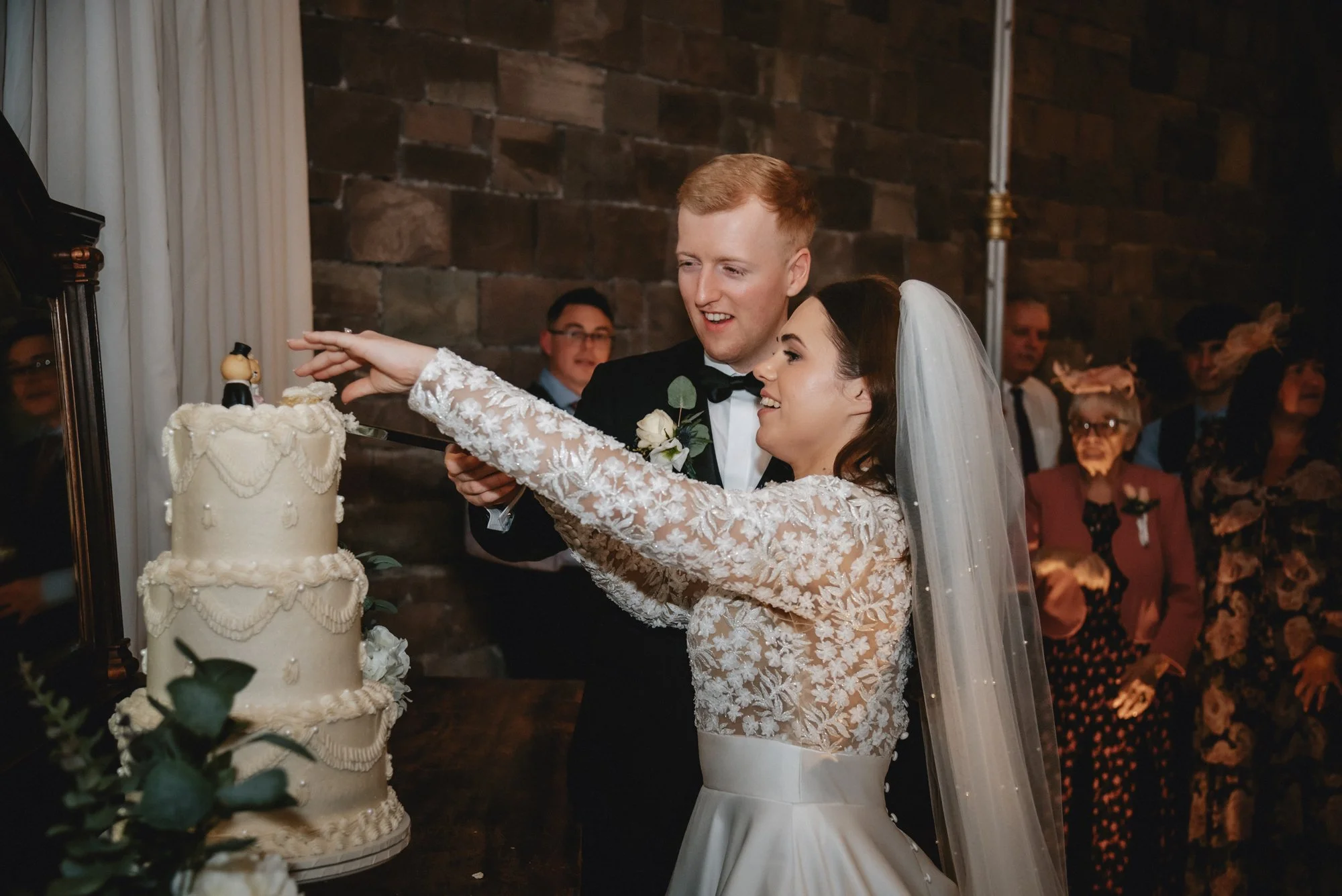 Bride and groom cutting wedding cake with figurine on top, surrounded by guests.