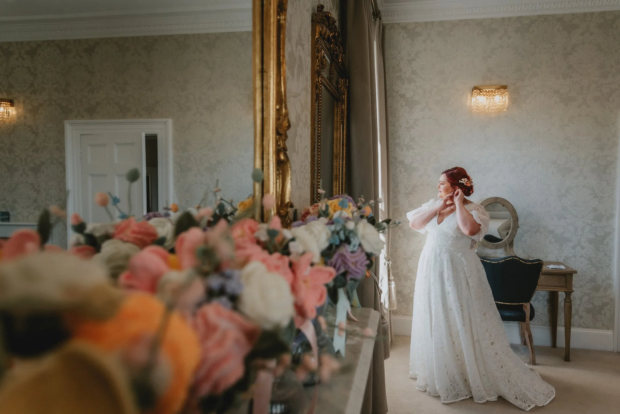 Brunette bride in a white lace wedding dress standing in a room, adjusting her earrings, with a mirror and elegant decor in the background, and a blurred bouquet of colorful flowers in the foreground.