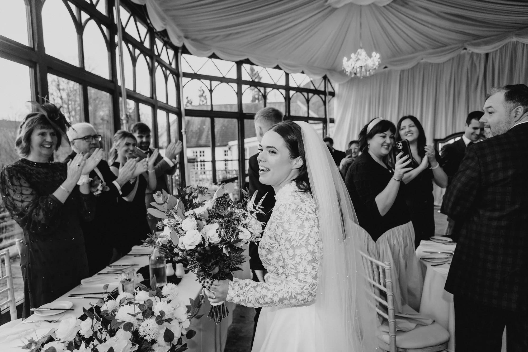 A bride holding a bouquet of flowers, smiling at her wedding reception, surrounded by guests clapping and smiling.