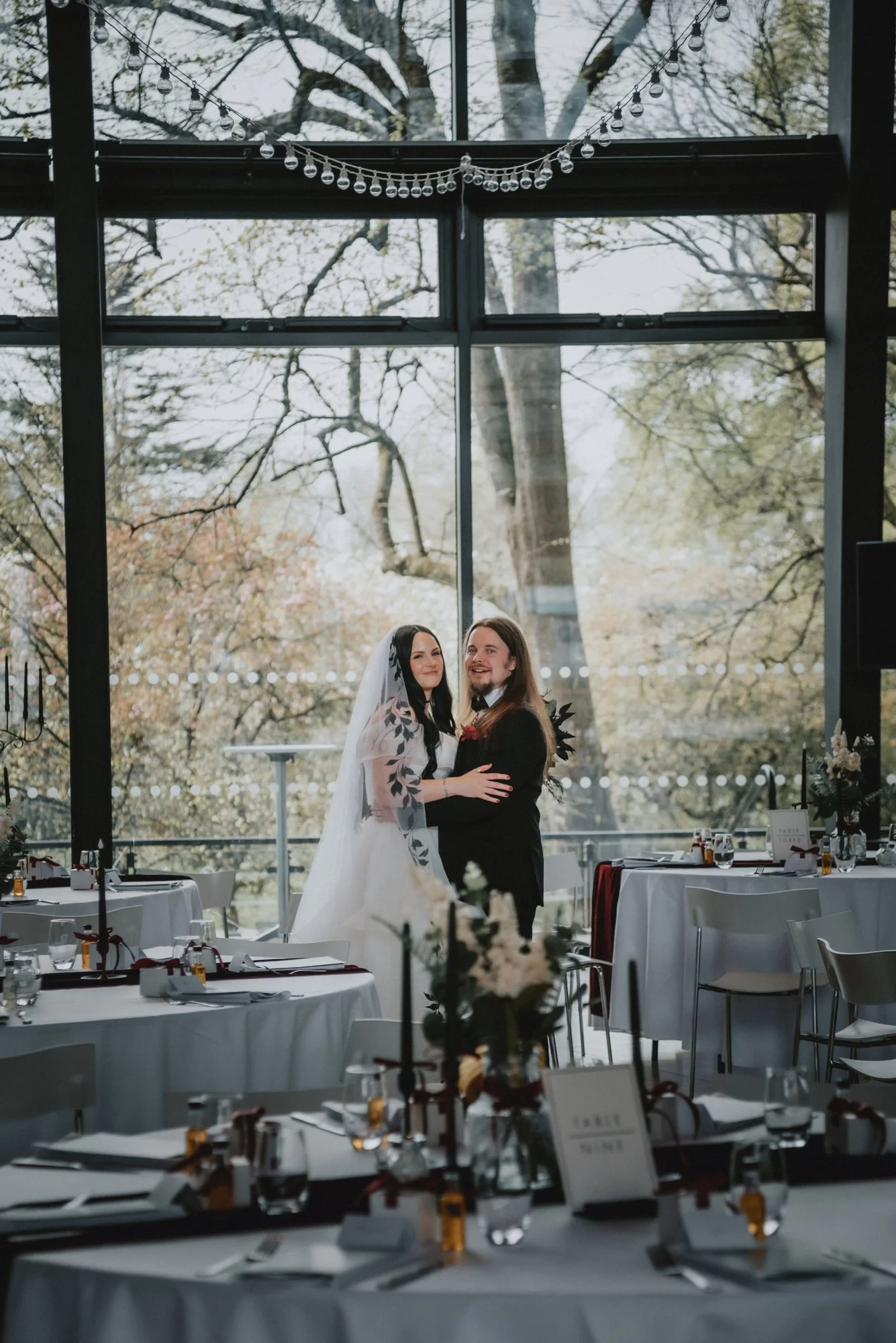 A bride and groom dancing inside a decorated venue with large windows and trees visible outside.