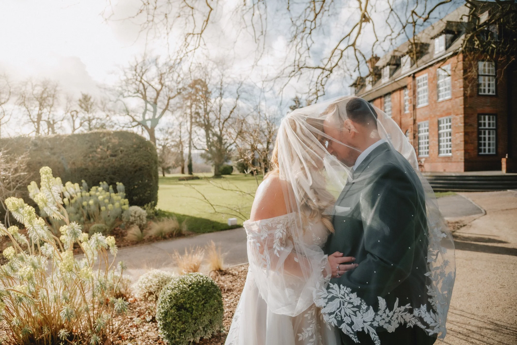 A bride and groom under a wedding veil sharing a kiss outdoors on a sunny day, with a brick building and leafless trees in the background.