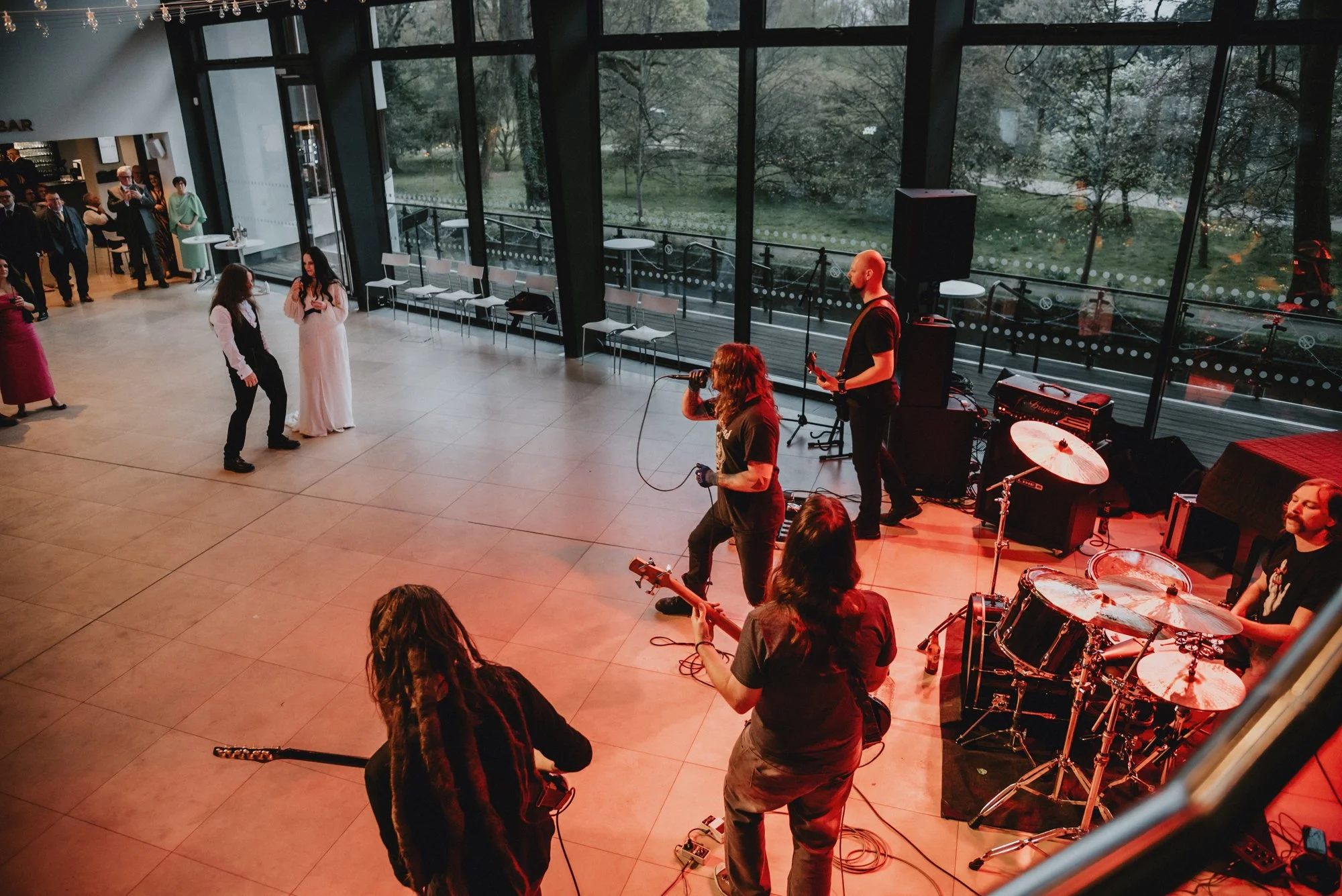 A band performs on stage at an indoor event with large windows overlooking a park. The band includes a guitarist and drummer, and the audience watches as two women dance near the stage.